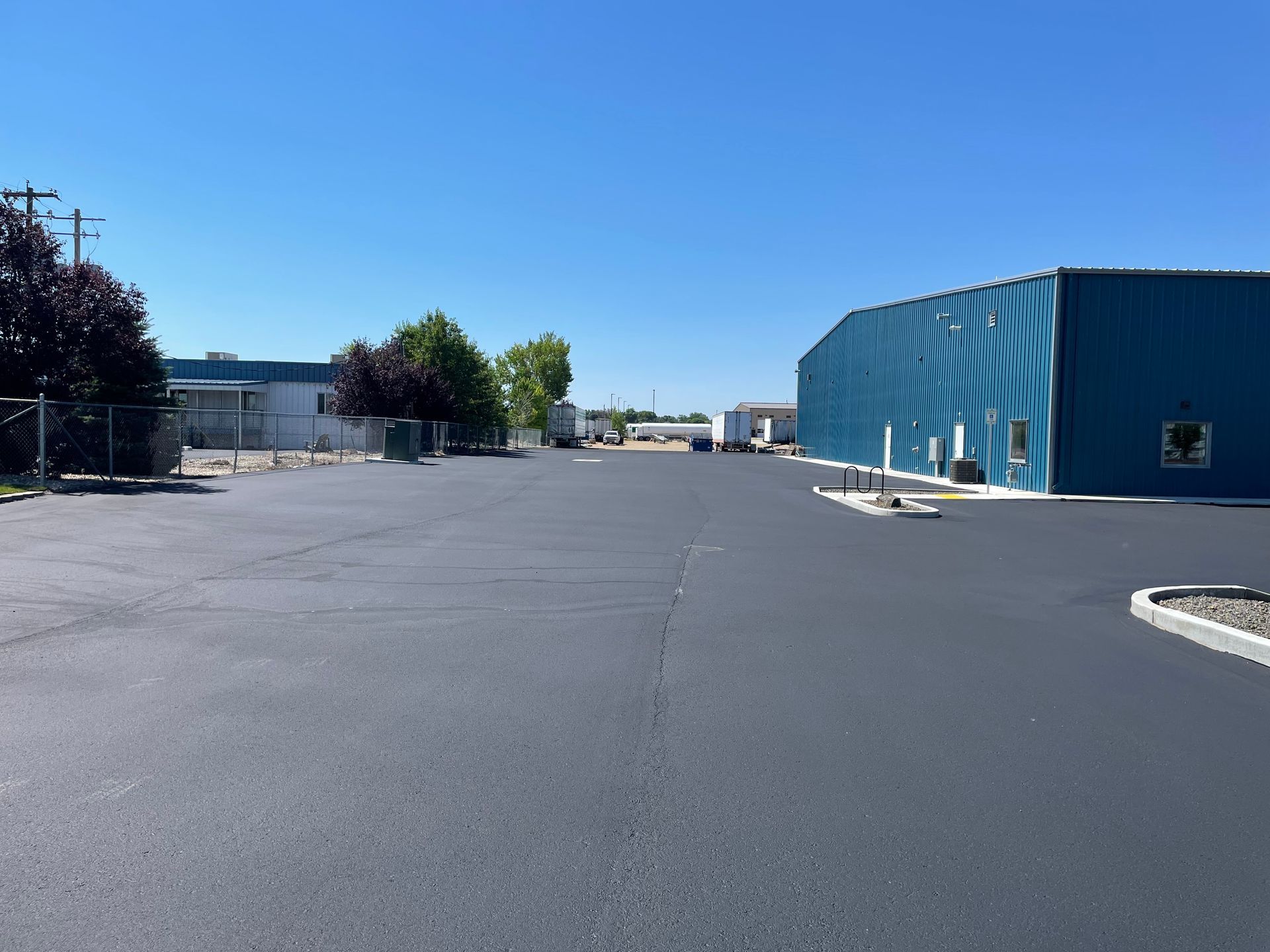 A view of a freshly paved, dark asphalt lot next to a long blue metal warehouse building under a clear blue sky.