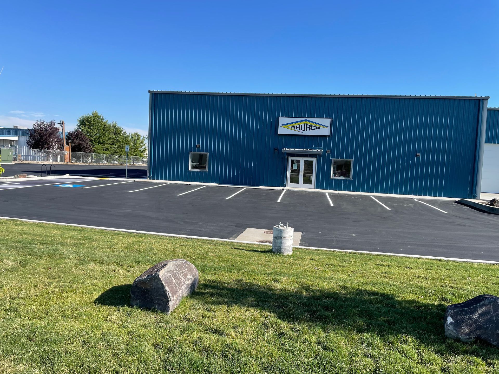 Blue commercial building with a white sign, entrance door, and a freshly paved parking lot under a clear blue sky.