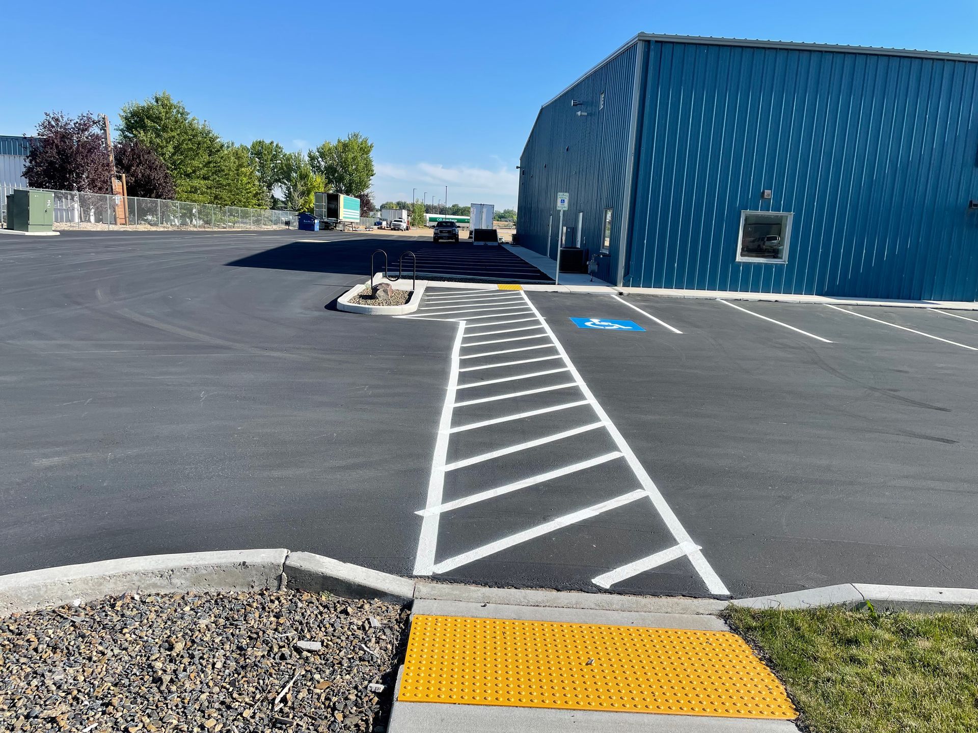 A paved parking lot featuring a crosswalk with yellow tactile paving leading toward a blue building and a handicap space.