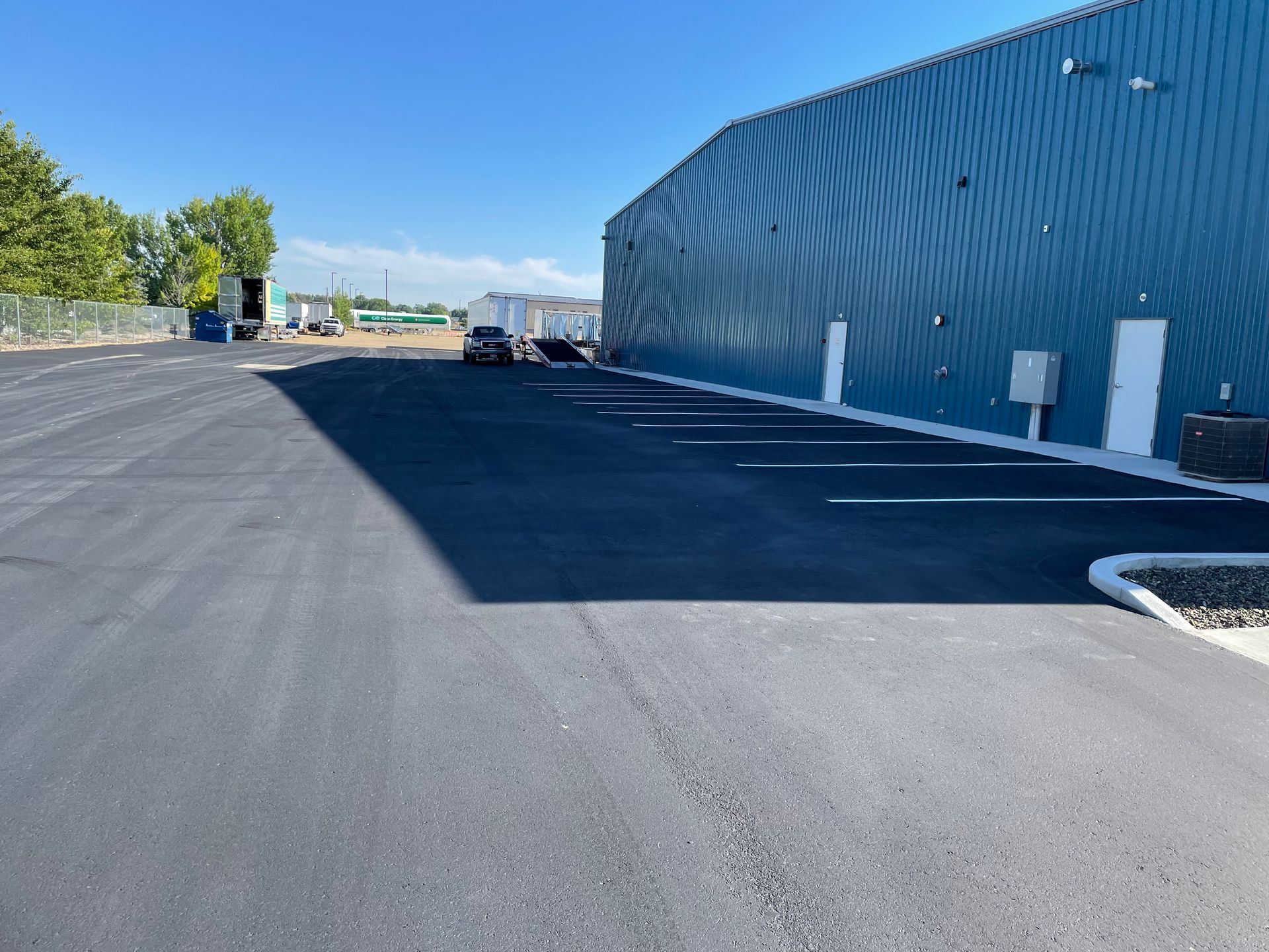 A newly paved asphalt parking lot beside a large blue metal industrial building under a clear blue sky.