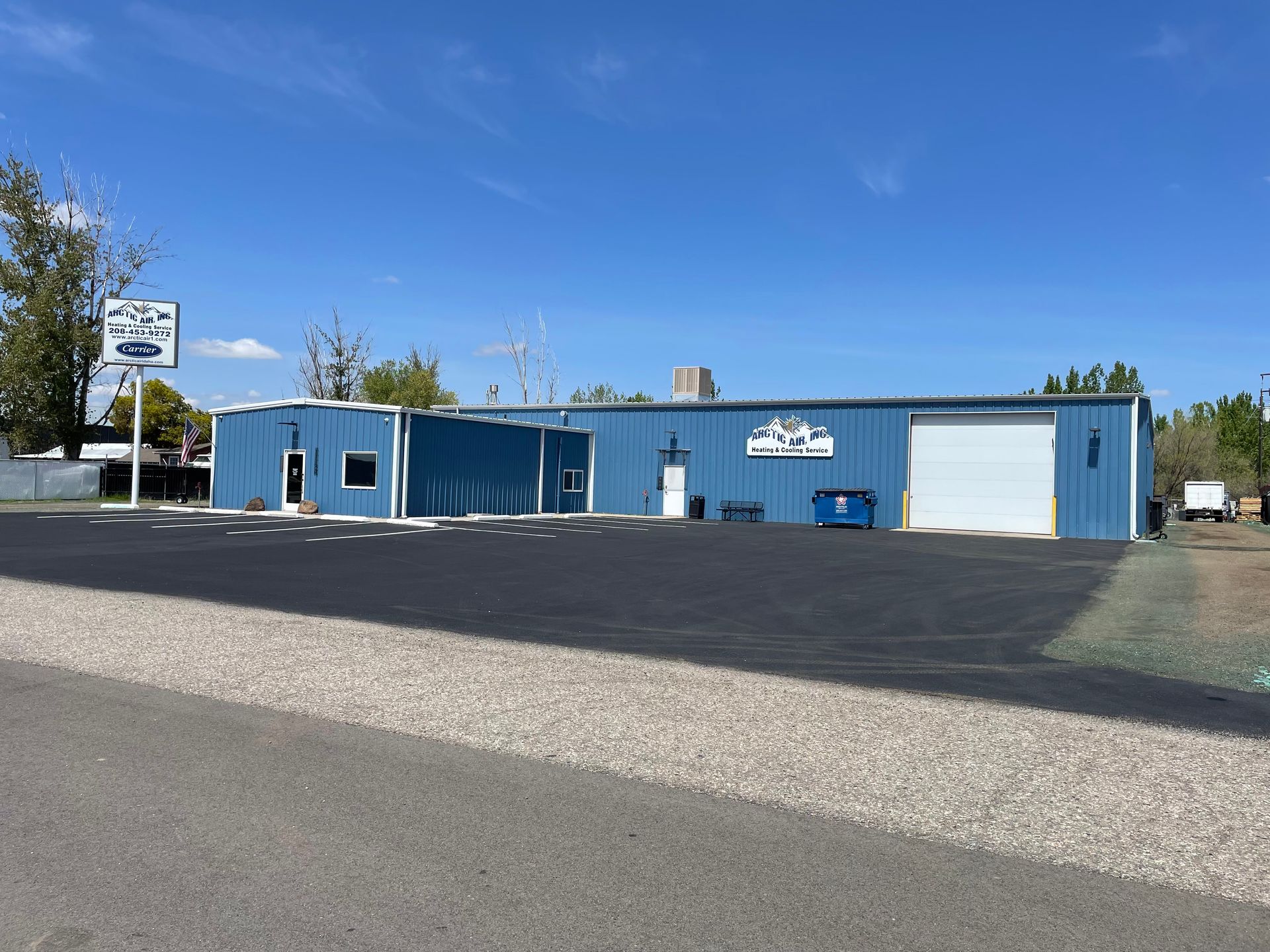 A blue commercial building with a large parking lot, a roll-up garage door, and a white sign under a clear blue sky.