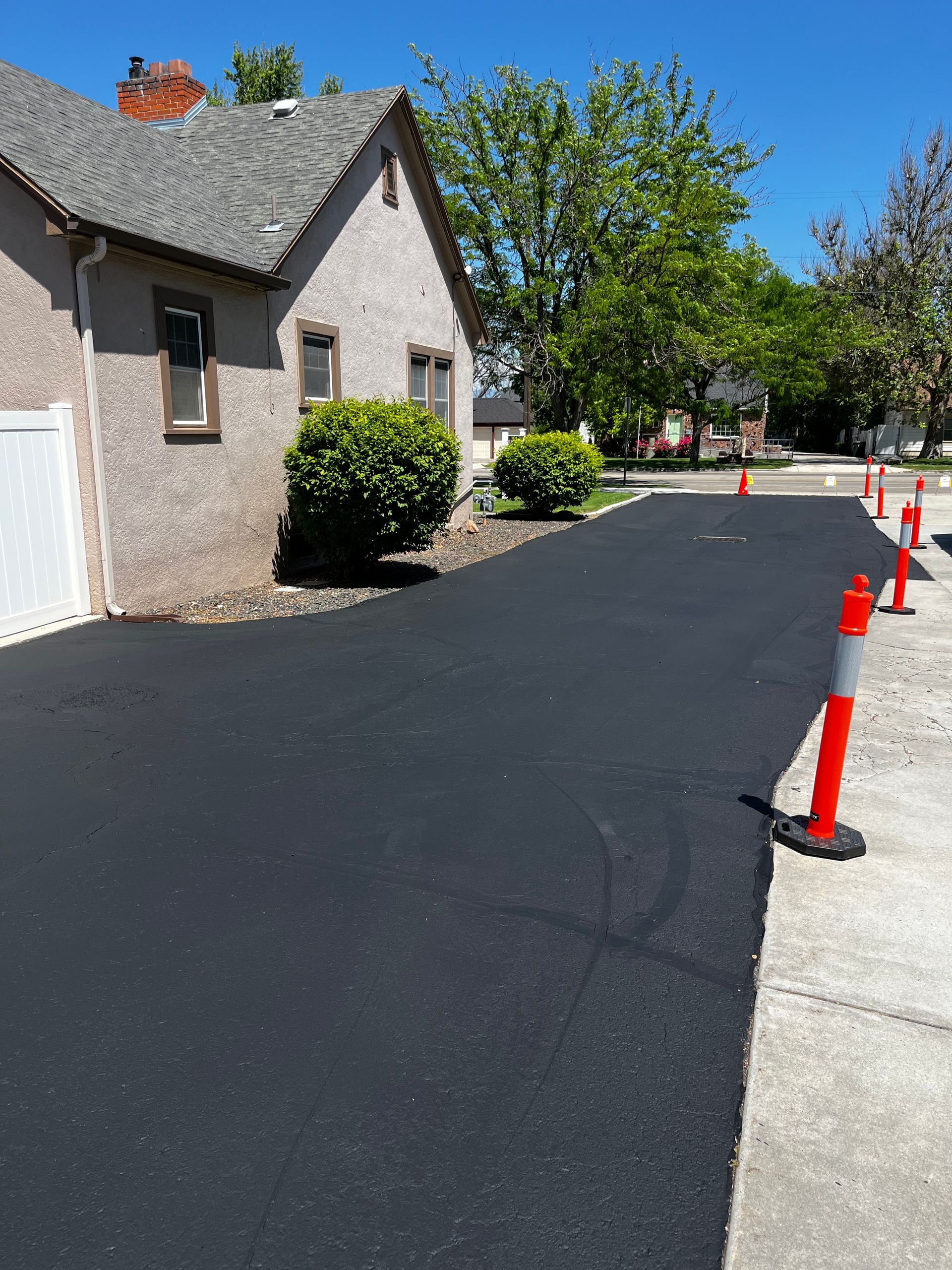 Freshly paved black asphalt driveway alongside a light-colored house with orange traffic cones lining the edge.