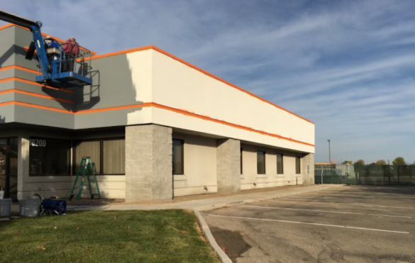 A boom lift operator paints a beige commercial building with orange accents, transitioning from gray to a lighter color.