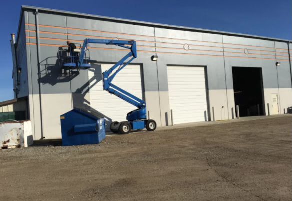 Two workers on a blue aerial lift platform perform maintenance on the upper exterior wall of a commercial building.
