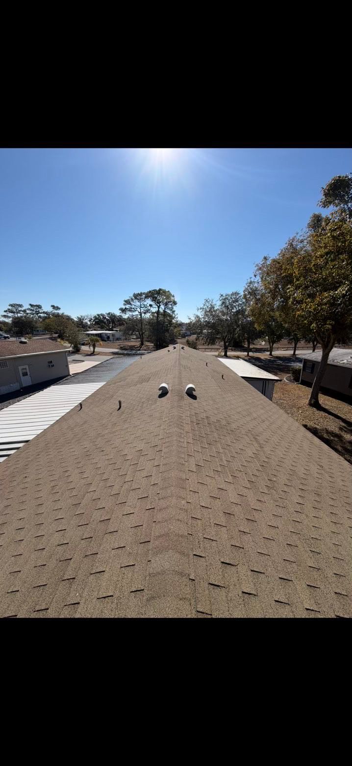 A view looking down the length of a tan, shingled roof under a clear blue sky, showing two small, white roof vents.