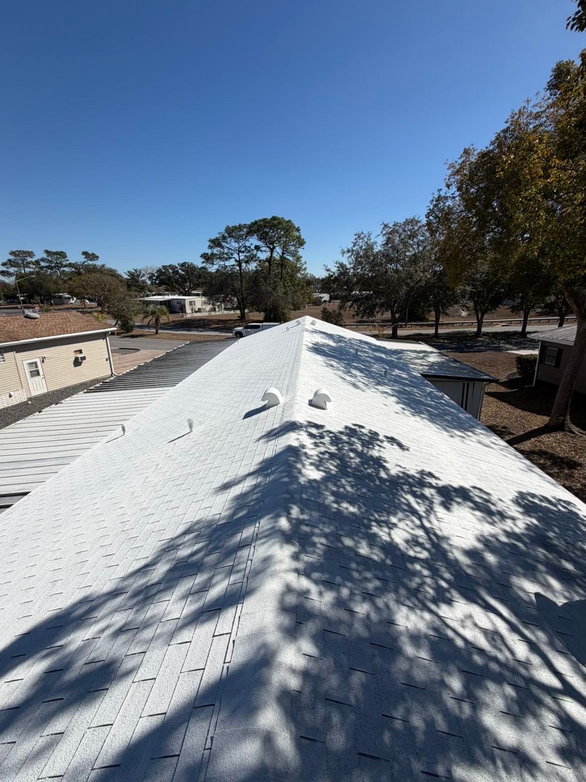 A view looking down the white-shingled roof ridge of a building, surrounded by trees and neighboring homes on a sunny day.