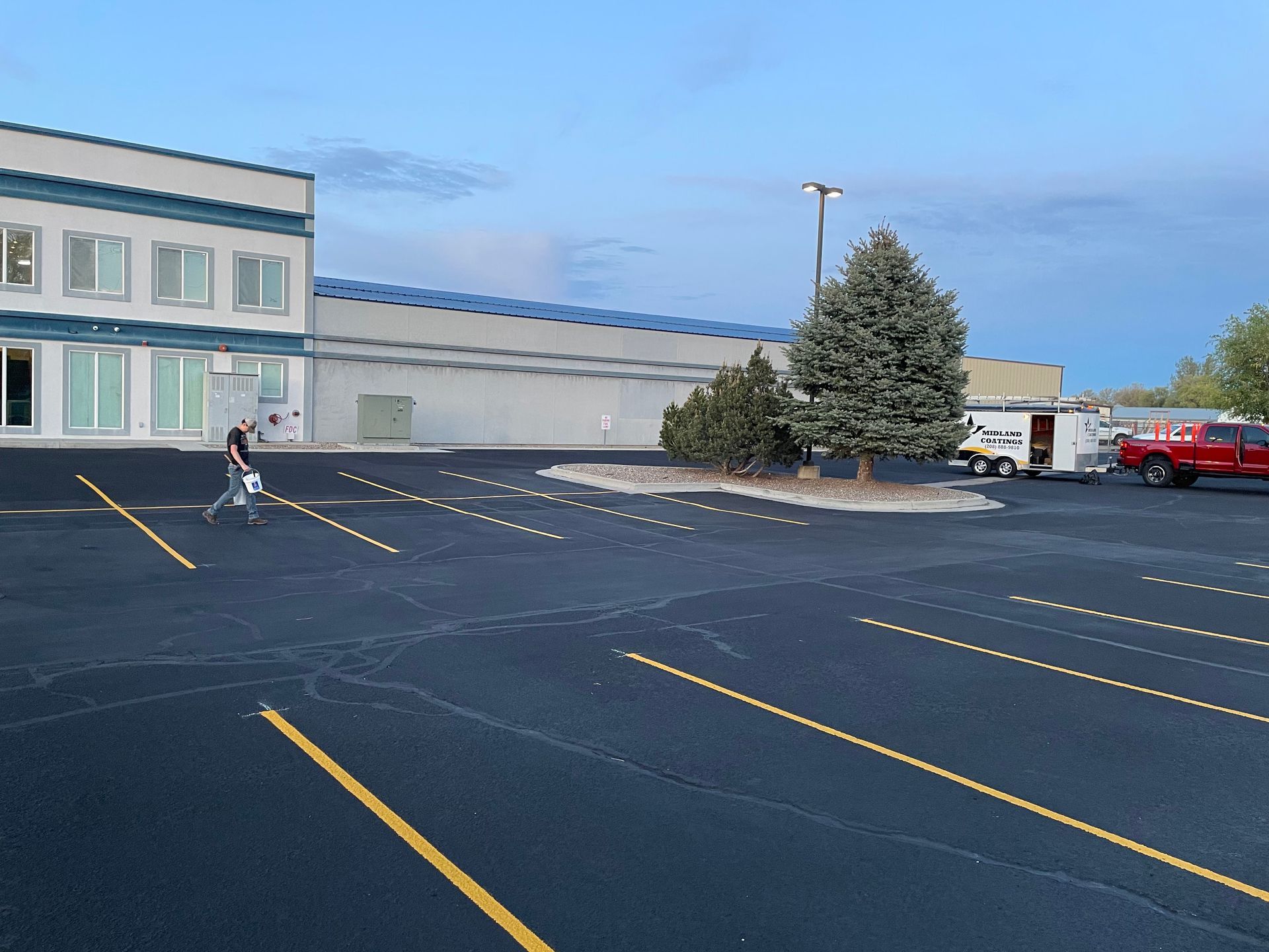 A person walks across a parking lot in front of a large, light-colored building with a blue roof at dusk.
