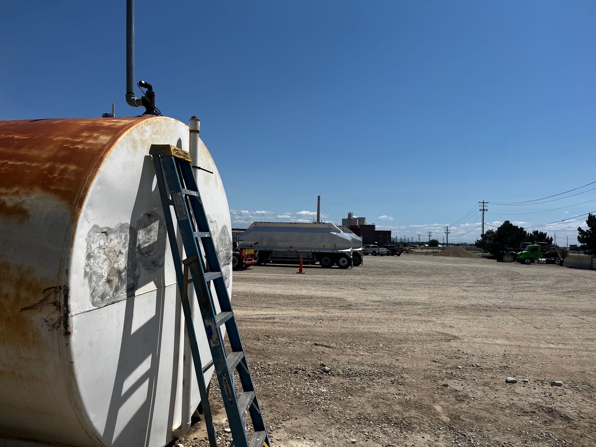 A metal tank with a leaning ladder stands in a gravel lot under a clear sky, with a fuel truck parked in the background.