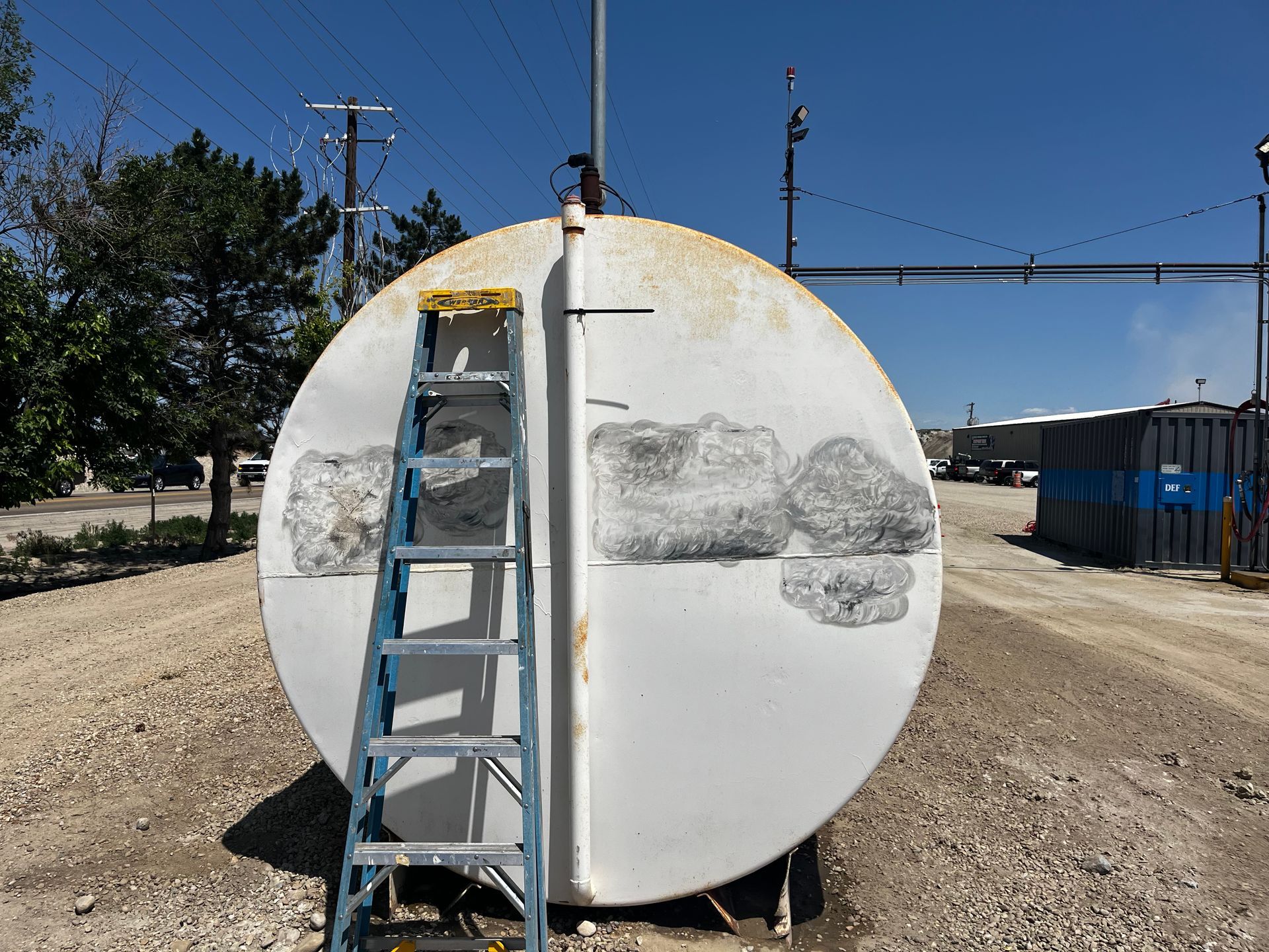 A tall ladder leans against a large, white circular storage tank outdoors on a gravel lot under a clear blue sky.