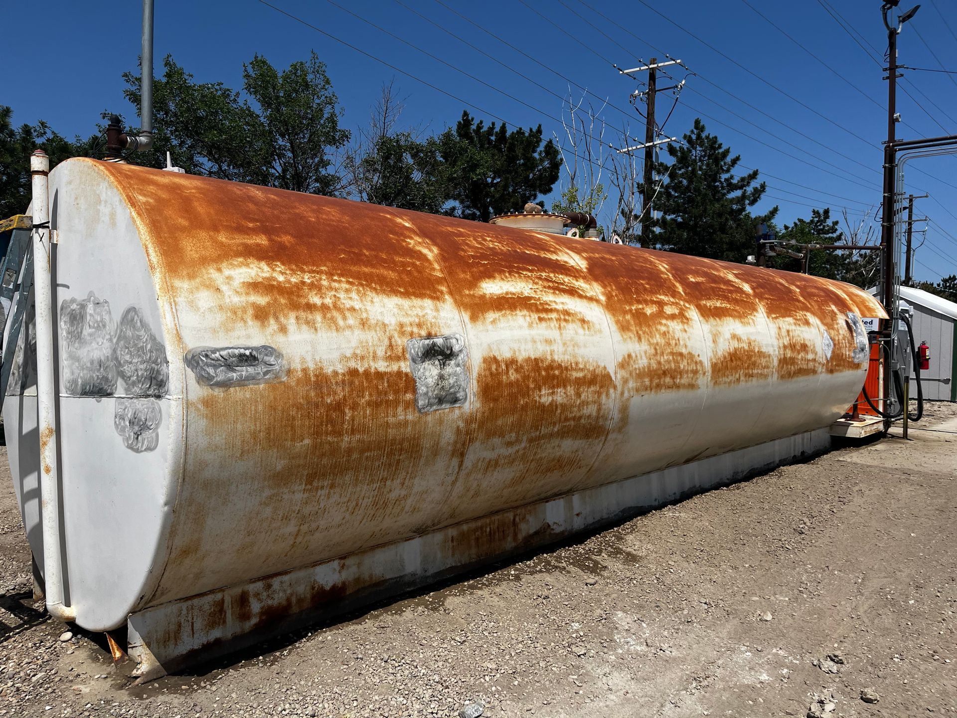 A large, cylindrical, horizontal metal storage tank with significant rust, sitting on gravel outdoors under a blue sky.