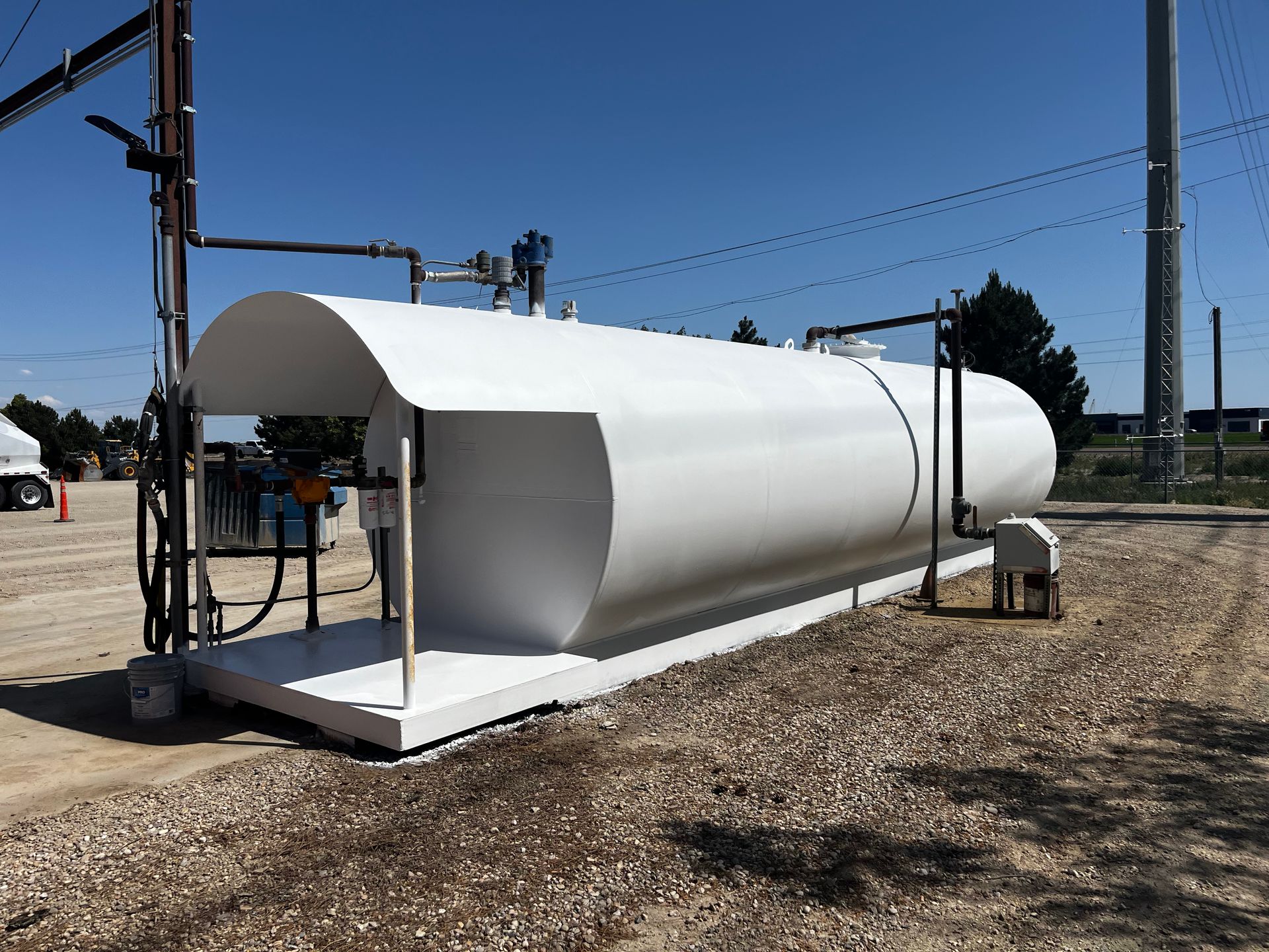 A white, horizontal fuel storage tank with a protective canopy sits on a gravel lot under a clear blue sky.