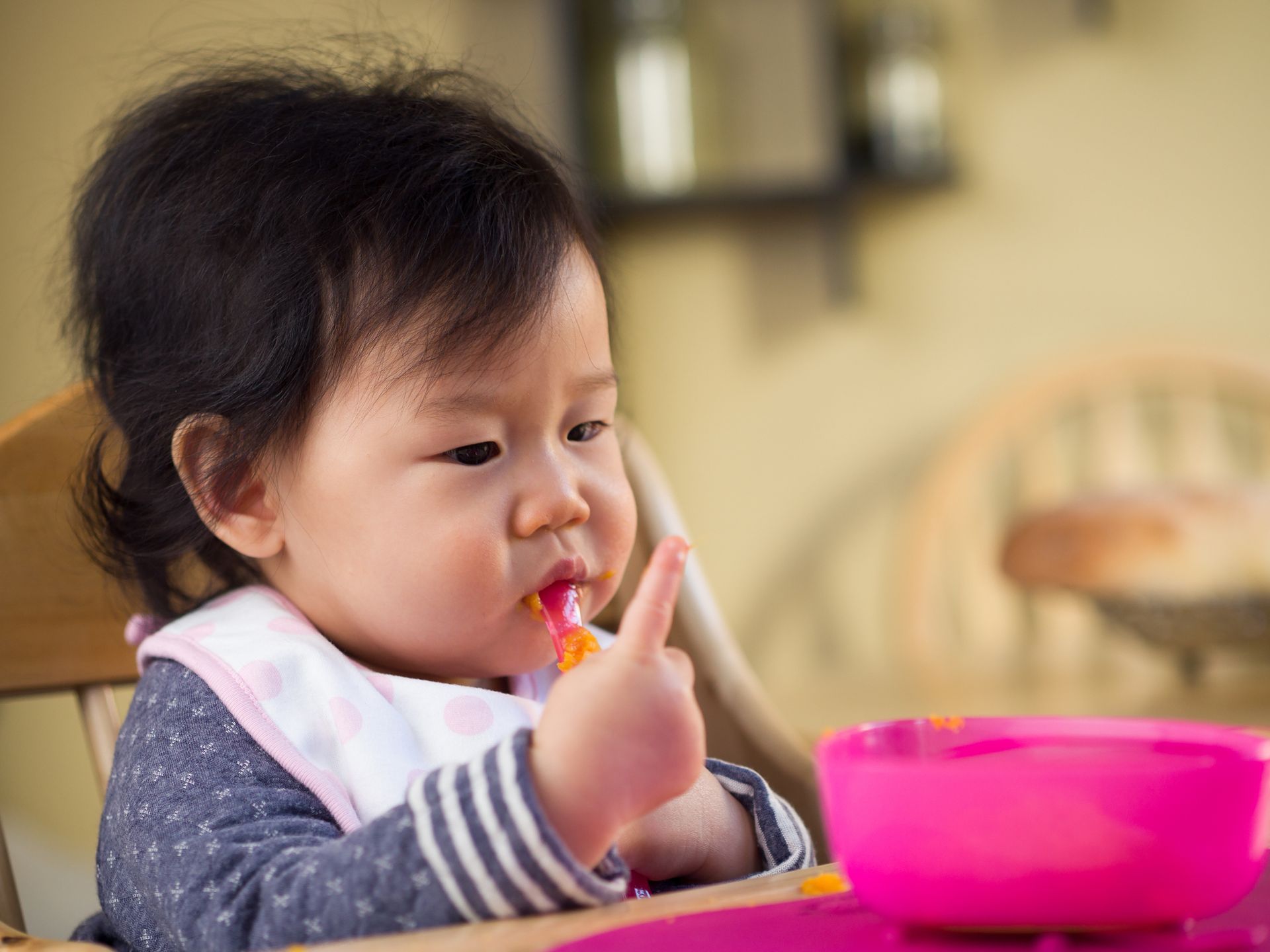 Baby eating from a pink bowl, food on finger and face, wearing bib, sitting in a high chair.
