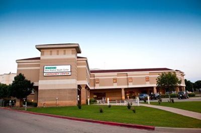 Hospital building with signage for emergency center; beige brick exterior, blue sky.