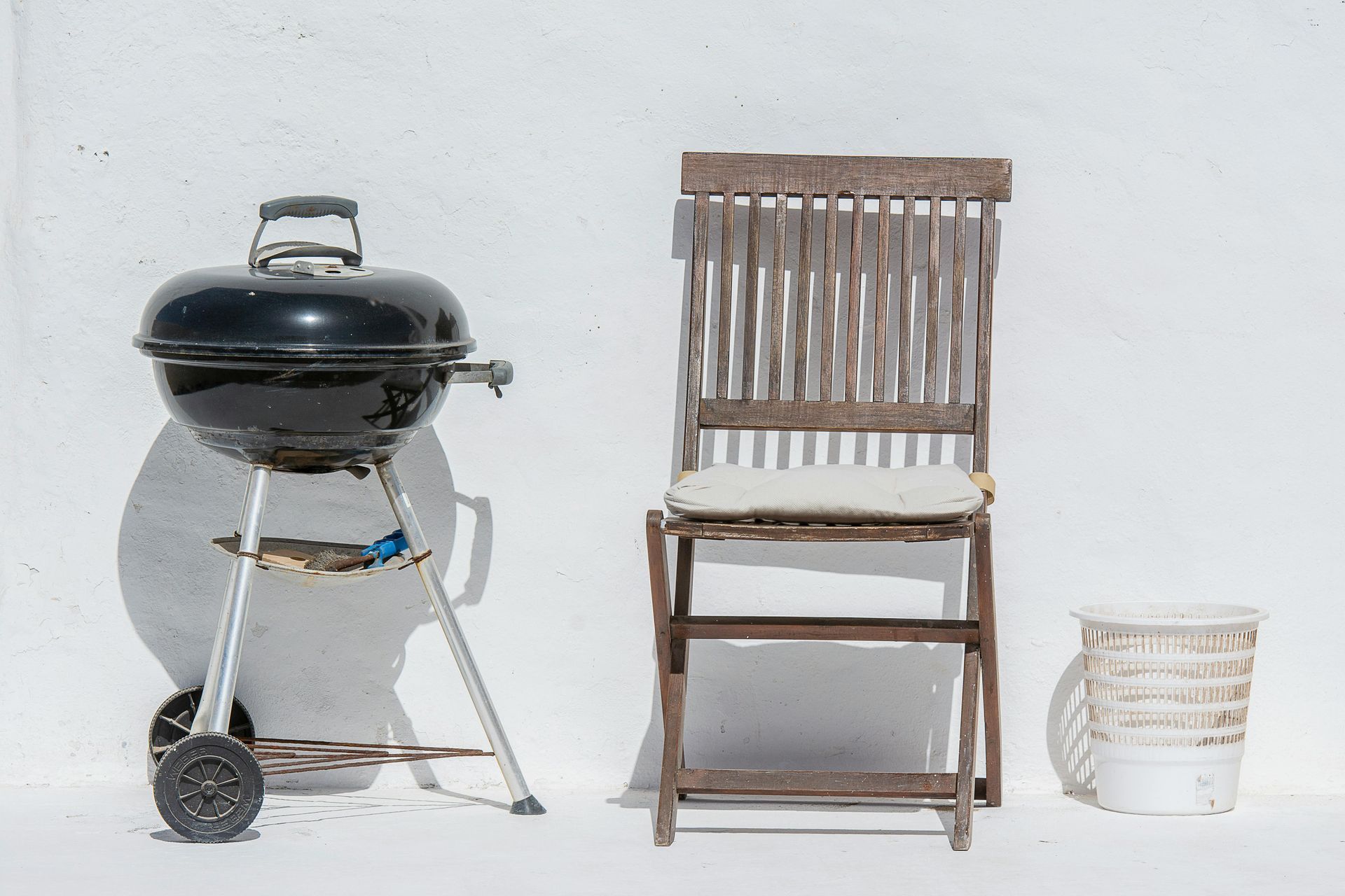 Black kettle grill, wooden chair, and white bucket on a plain background.