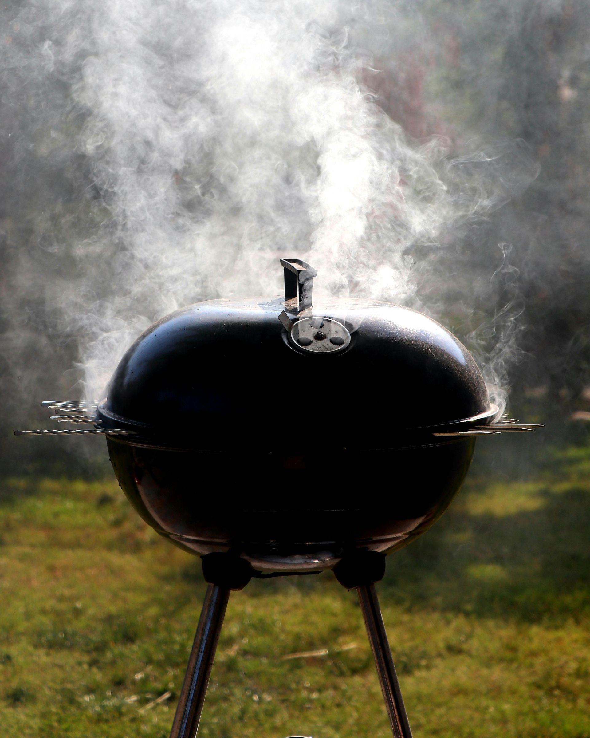 Black charcoal grill on tripod with smoke rising in a grassy yard