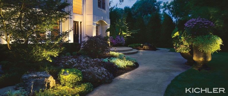 Illuminated garden pathway beside a house at dusk, with landscape lighting highlighting shrubs and trees