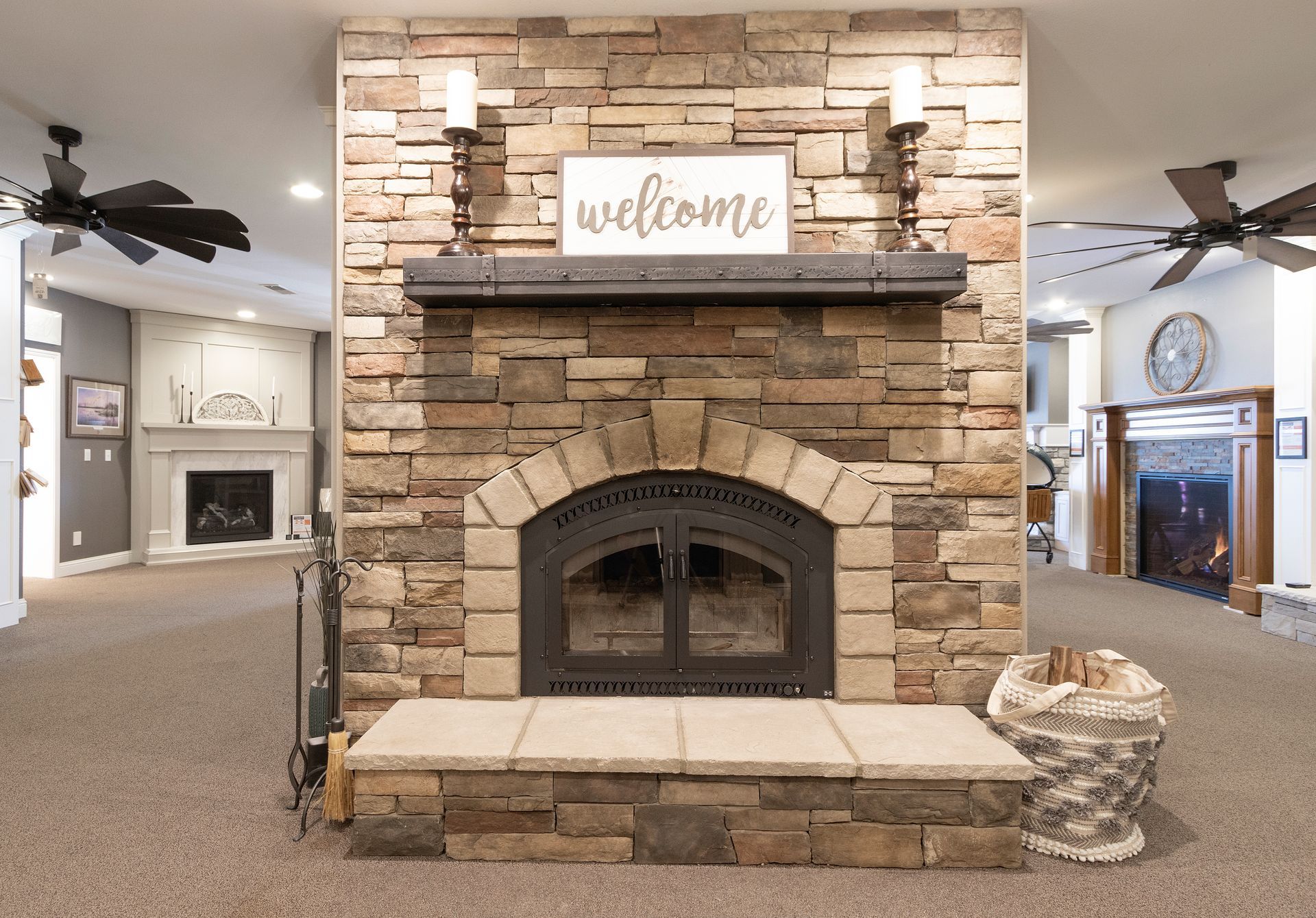 Stone fireplace with a “welcome” sign in a carpeted living room, with ceiling fans on both sides