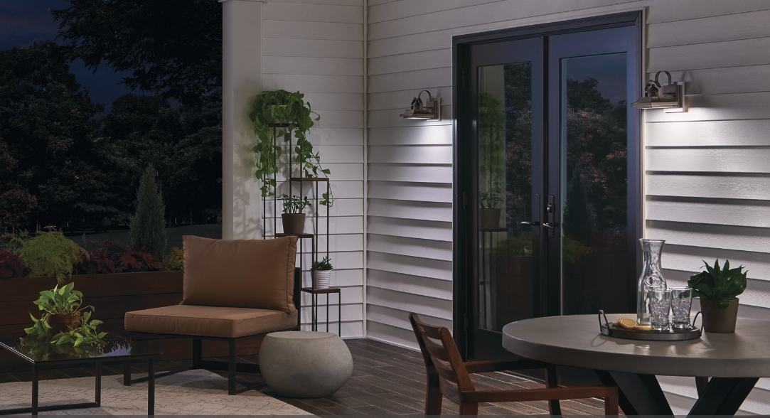 Lit patio with wicker chair, round table, potted plants, and glass doors beside a house at dusk