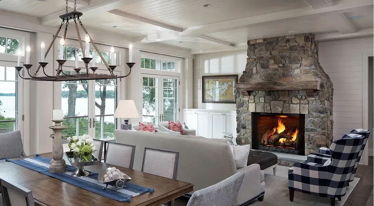 Bright modern living-dining room with stone fireplace, chandelier, and large windows overlooking greenery