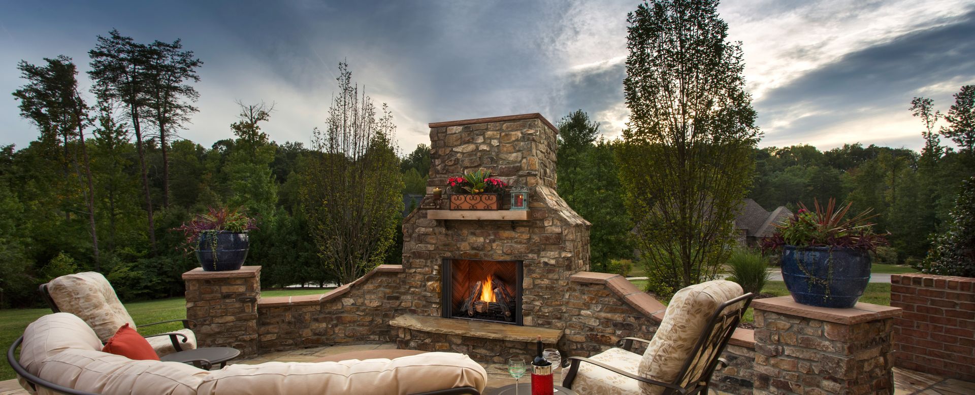 Stone patio with cushioned chairs around a lit outdoor fireplace at sunset, overlooking trees and hills