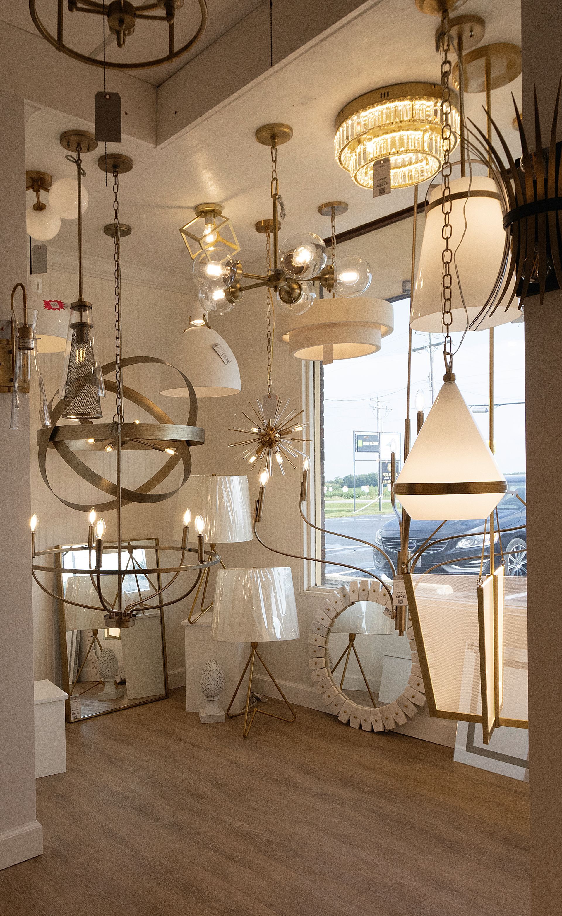Assorted hanging lamps and chandeliers displayed in a bright showroom with a window and wood floor