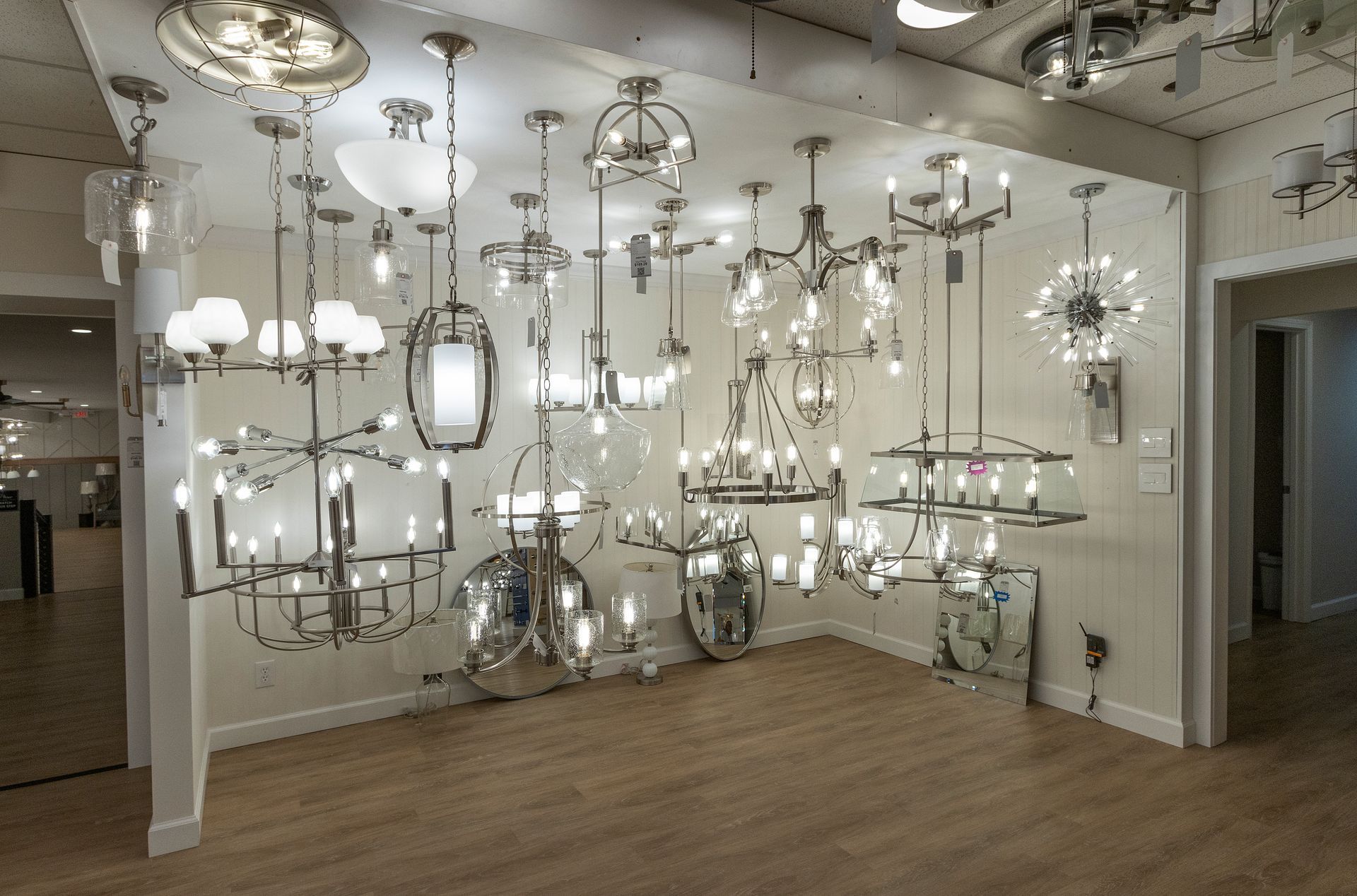 Showroom of assorted chrome and glass chandeliers and pendant lights on display with wooden floor and white walls