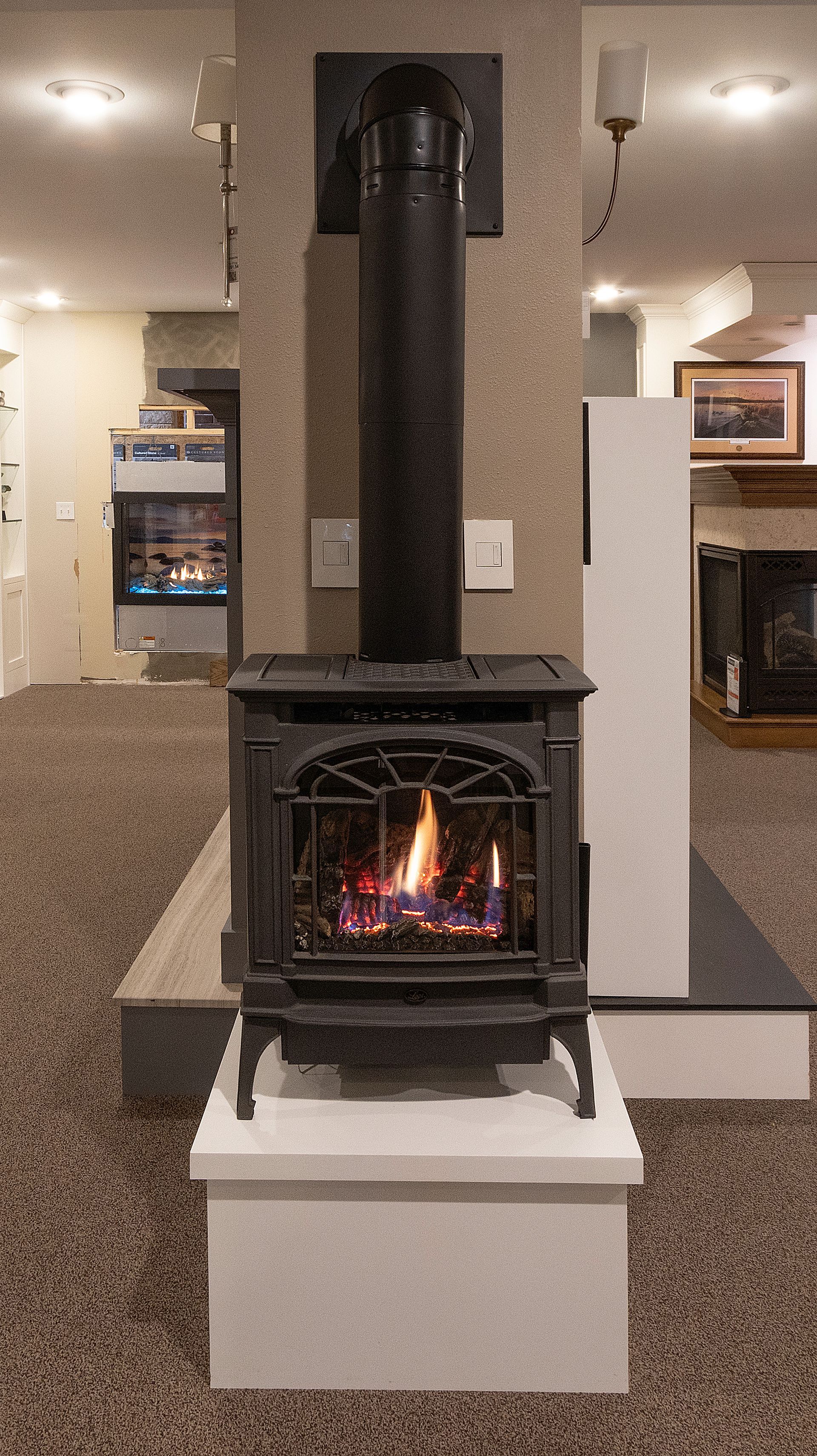 Modern black wood stove with chimney in a showroom, flames visible inside, on a white pedestal.