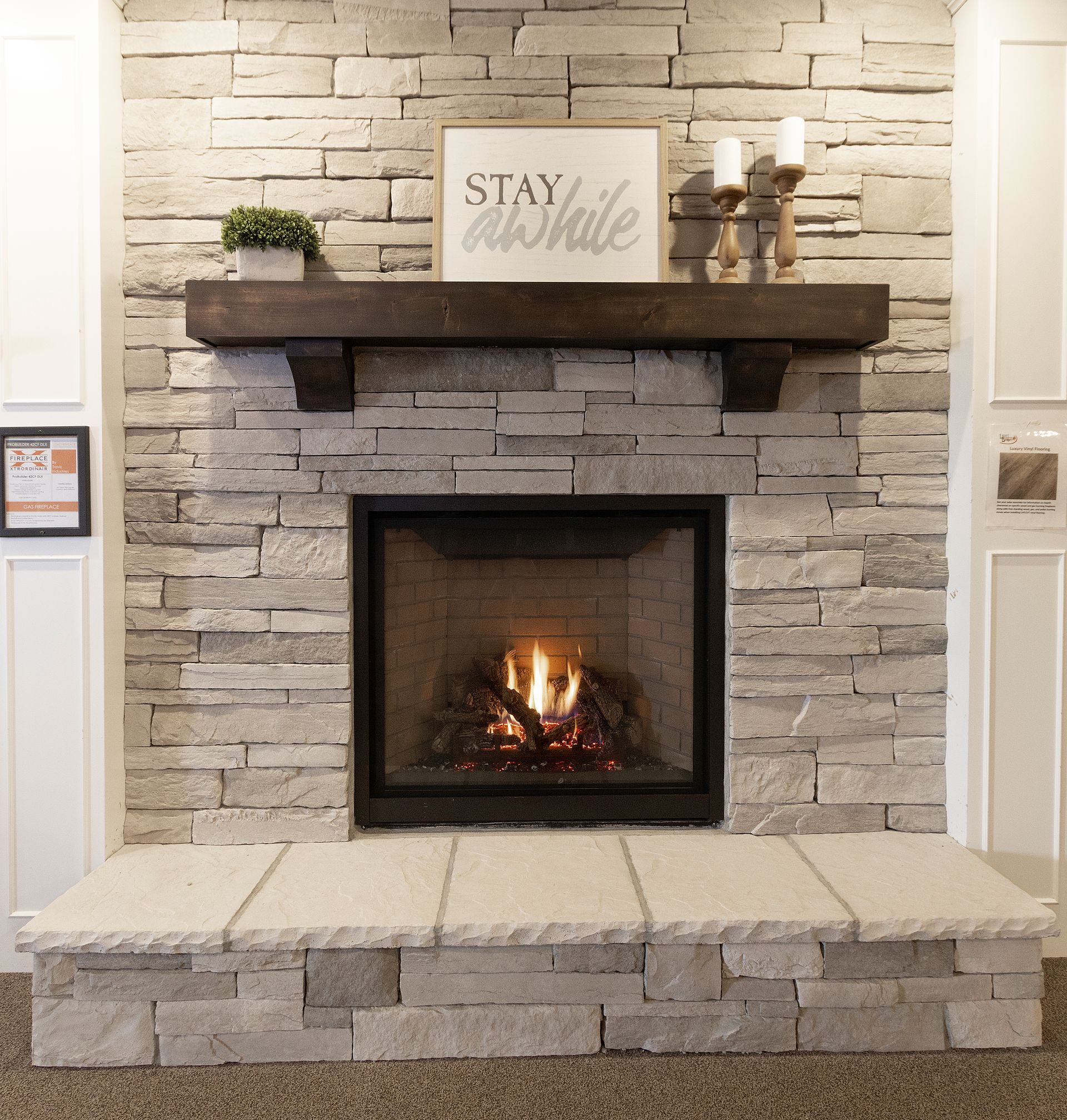 Stone fireplace with a lit fire, dark mantel, and “stay awhile” sign above a hearth bench.
