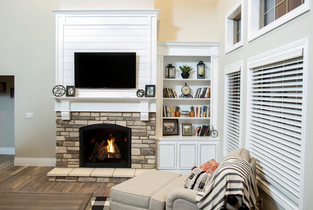 Bright living room with a stone fireplace, wall-mounted TV, white shelves, and a striped chair by the window.