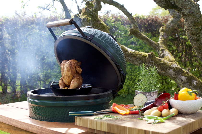 Steak grilling on a green kamado grill beside fresh vegetables on a wooden outdoor table