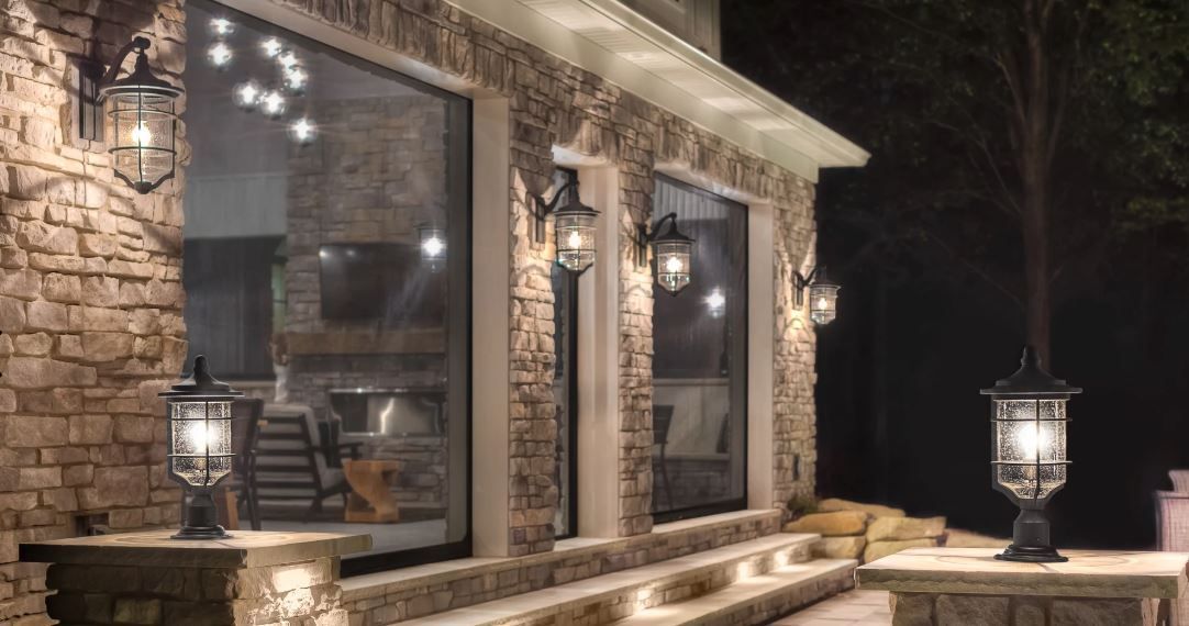 Stone patio with lit lanterns and a large window beside a modern rustic house at night