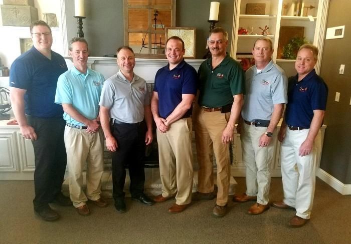 Seven men posing indoors in a living room, standing side by side and smiling at the camera.