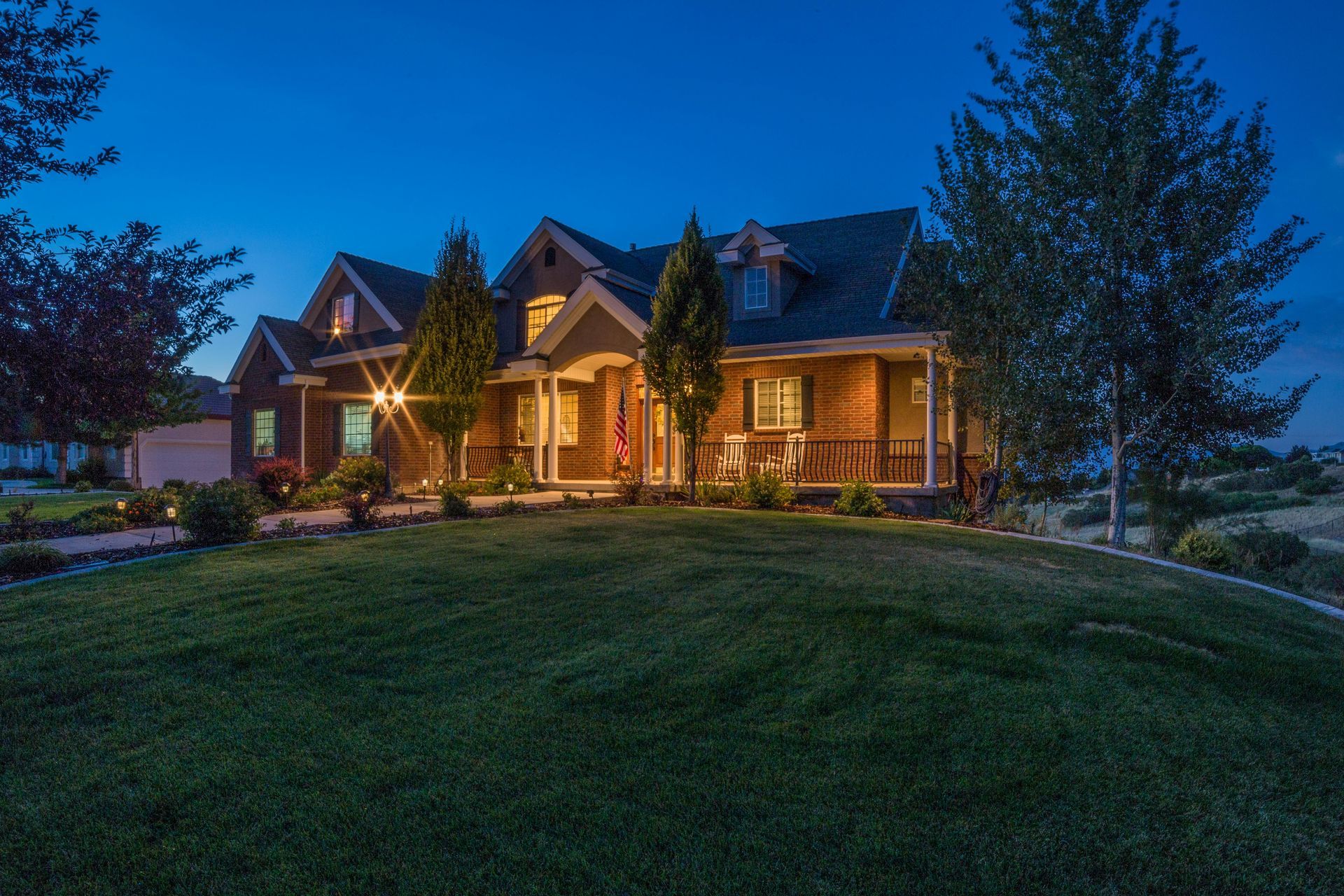 A warmly lit house at dusk with a front porch, trees, and a dark green lawn.