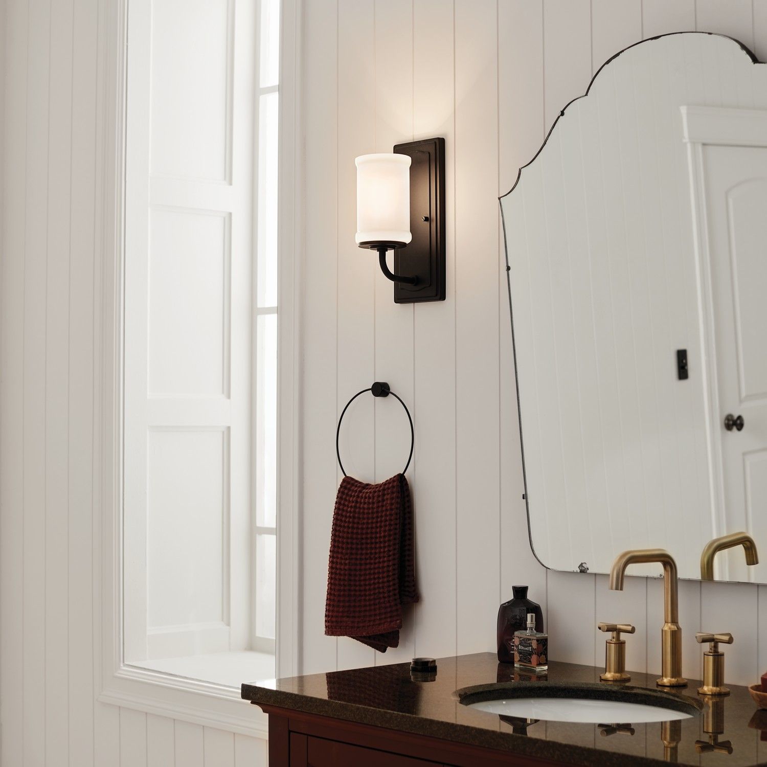 Bright bathroom with wall sconce, dark vanity, gold faucet, and scalloped mirror