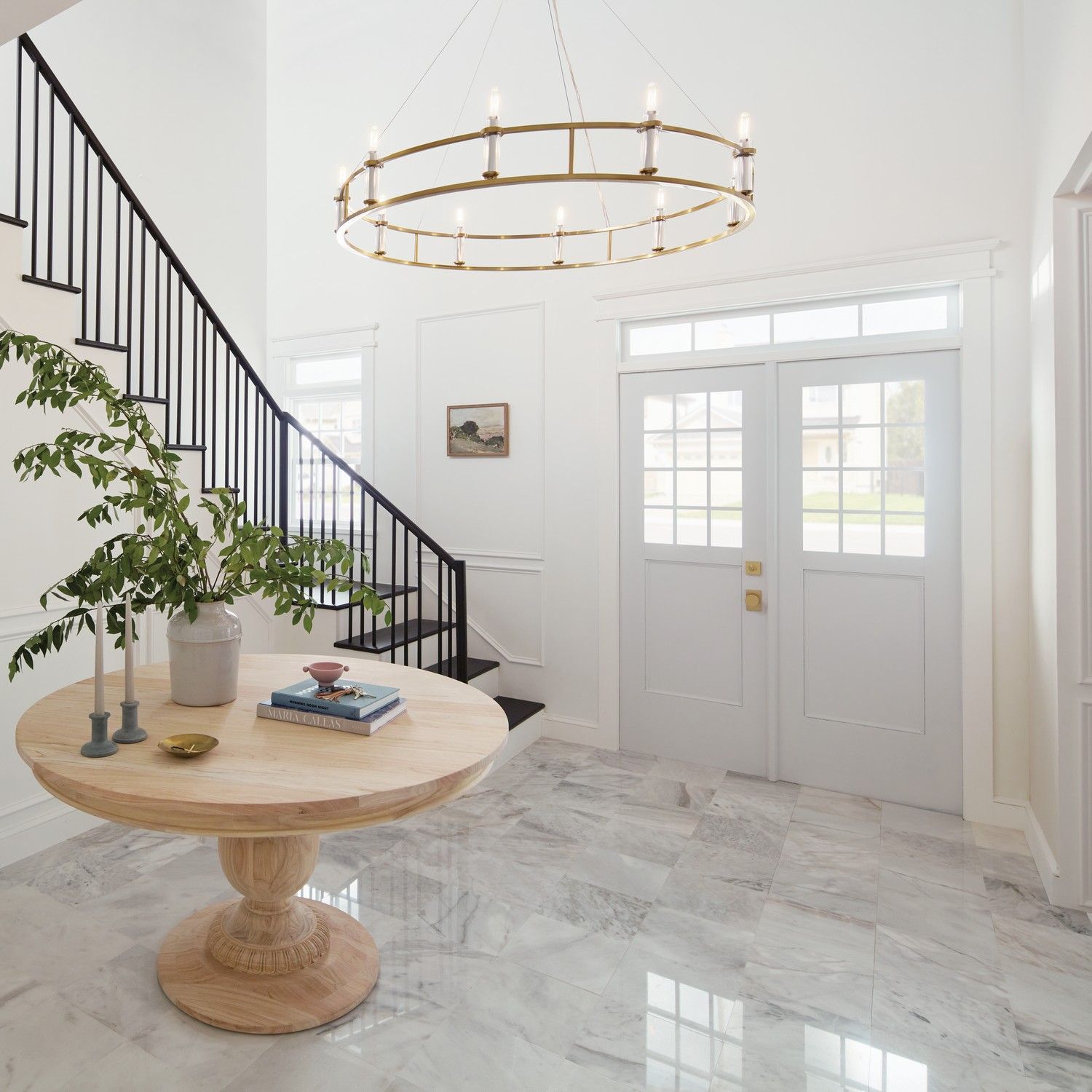 Bright foyer with marble floor, round wooden table, black staircase, and gold chandelier
