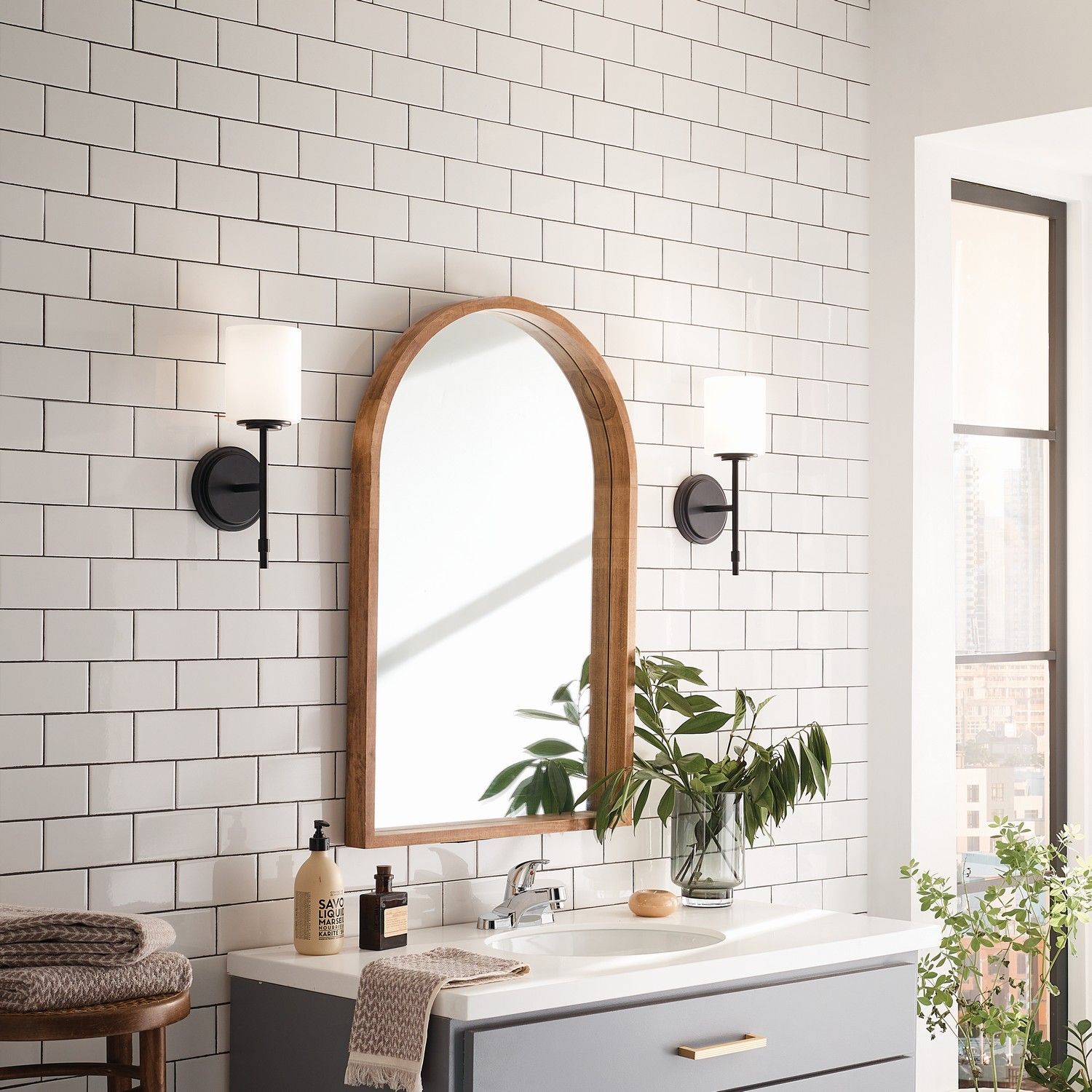 Bright bathroom vanity with arched mirror, wall sconces, white tile, and potted plants