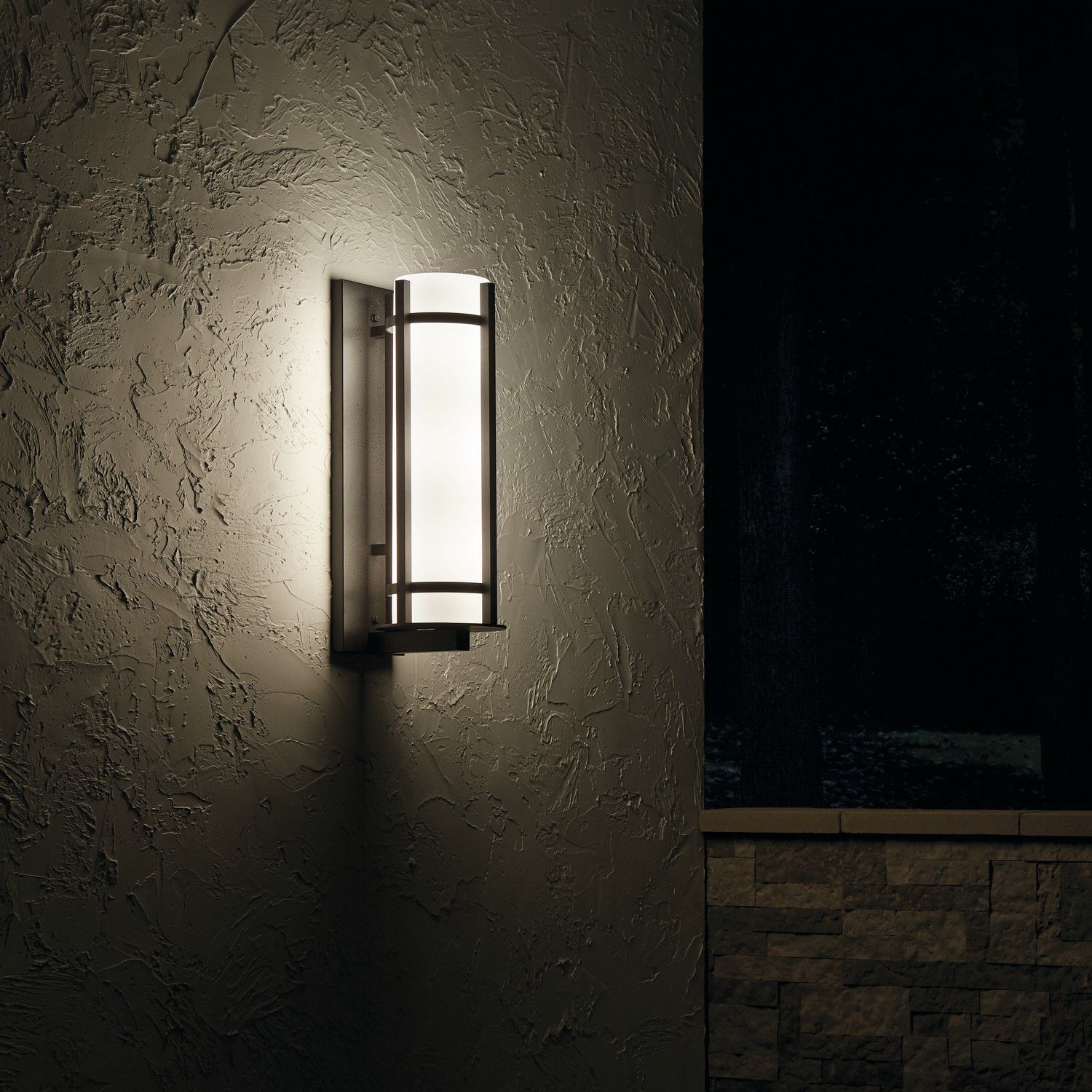 Wall-mounted lantern casting warm light on a textured exterior wall beside a dark window.