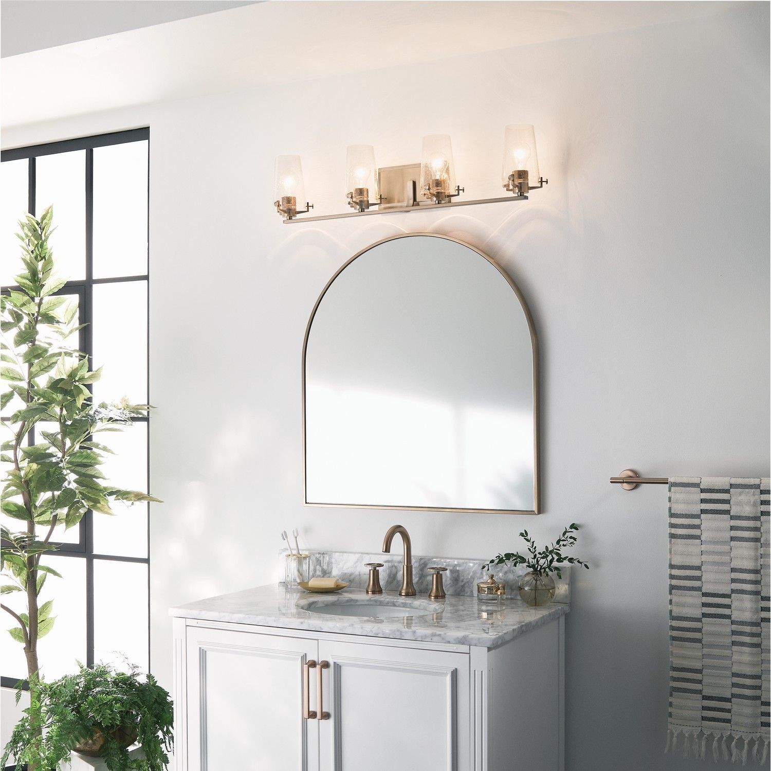 Modern white bathroom vanity with arched mirror, brass sconces, and plant by a window