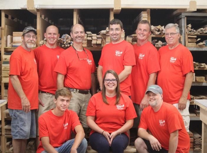 Group of people in red shirts posing together in a workshop with stacked wooden materials behind them