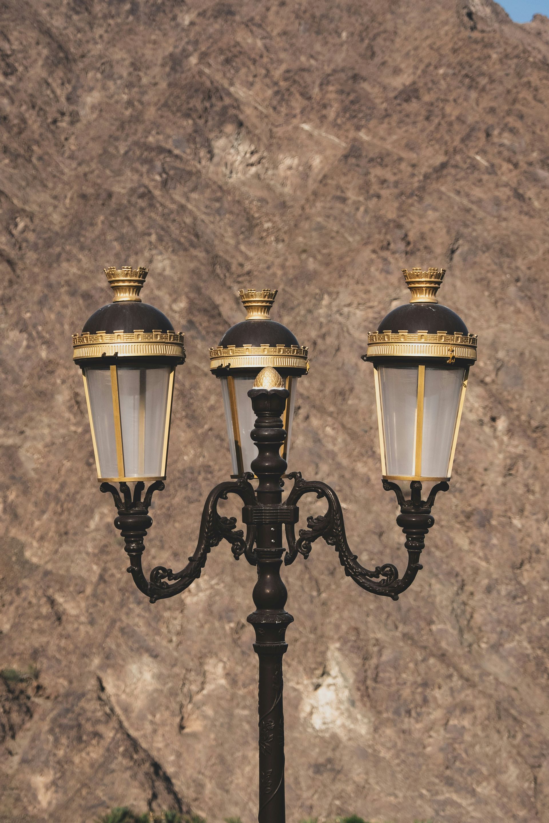 Ornate black and gold streetlamp with three lanterns against a rocky hillside.