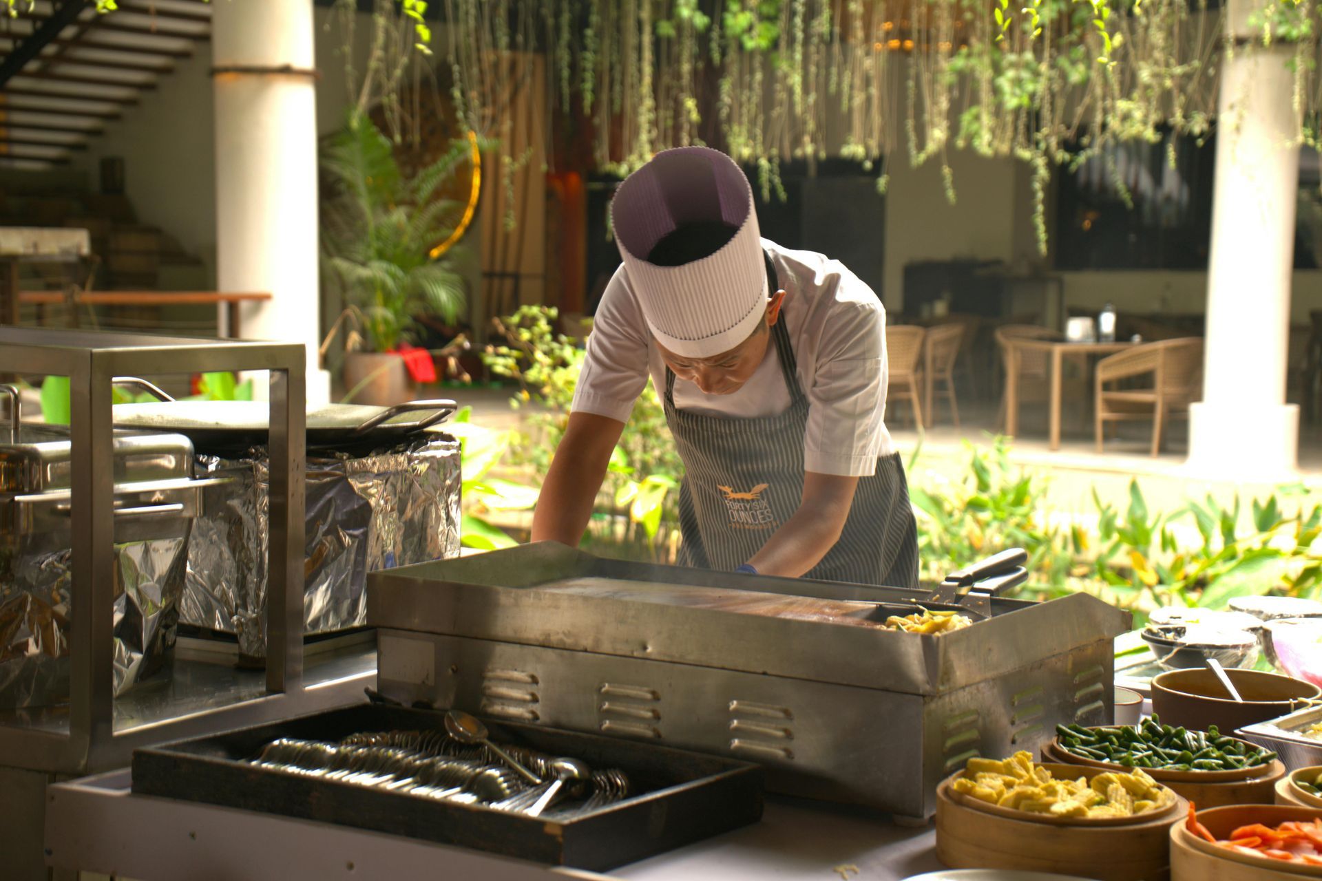 Chef grilling food at an outdoor market stall under hanging greenery