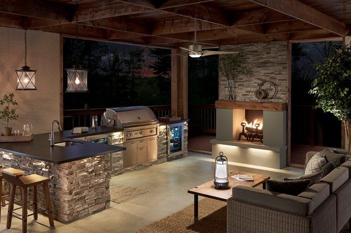 Covered outdoor patio with stone barbecue, wicker seating, fireplace, and warm lighting at dusk