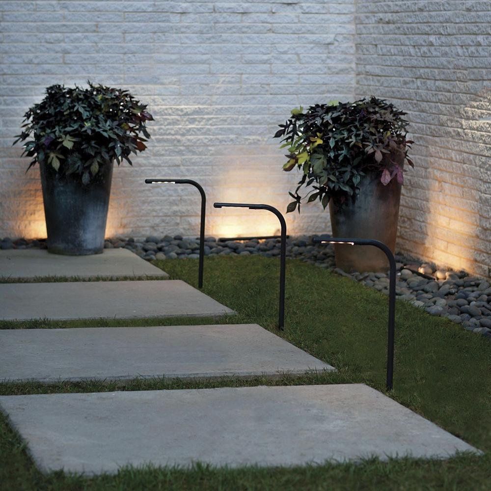 Outdoor walkway with concrete stepping stones, black handrails, potted plants, and wall lighting along a brick wall