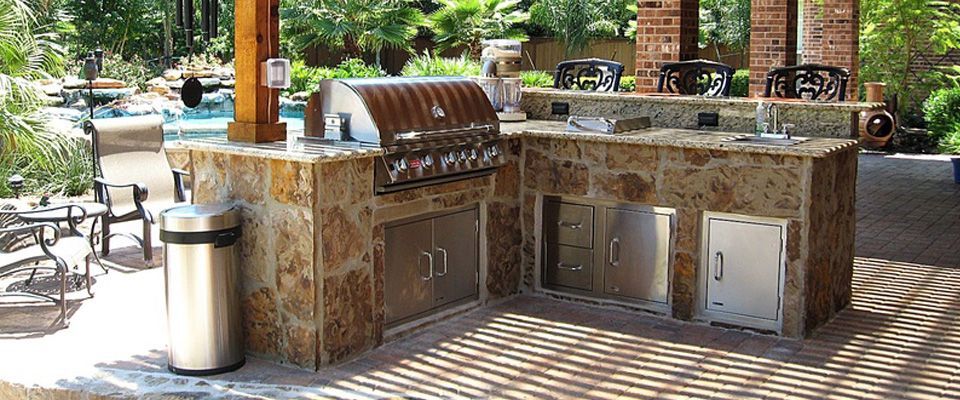 Outdoor stone grill and sink area beside a park path with a trash can and picnic seating