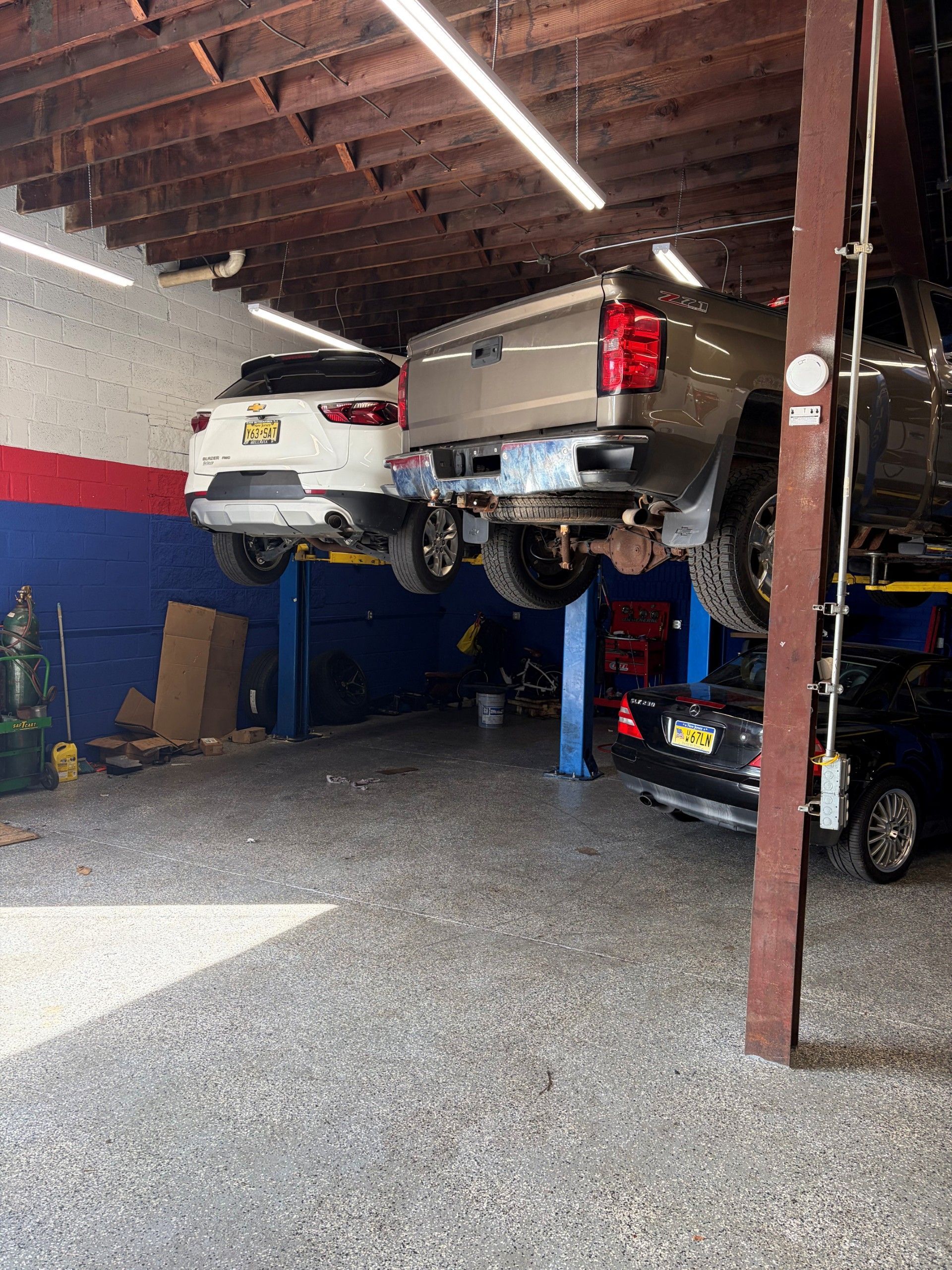 Two vehicles lifted in an auto repair shop, with a red, white, and blue wall and a blue car at right.