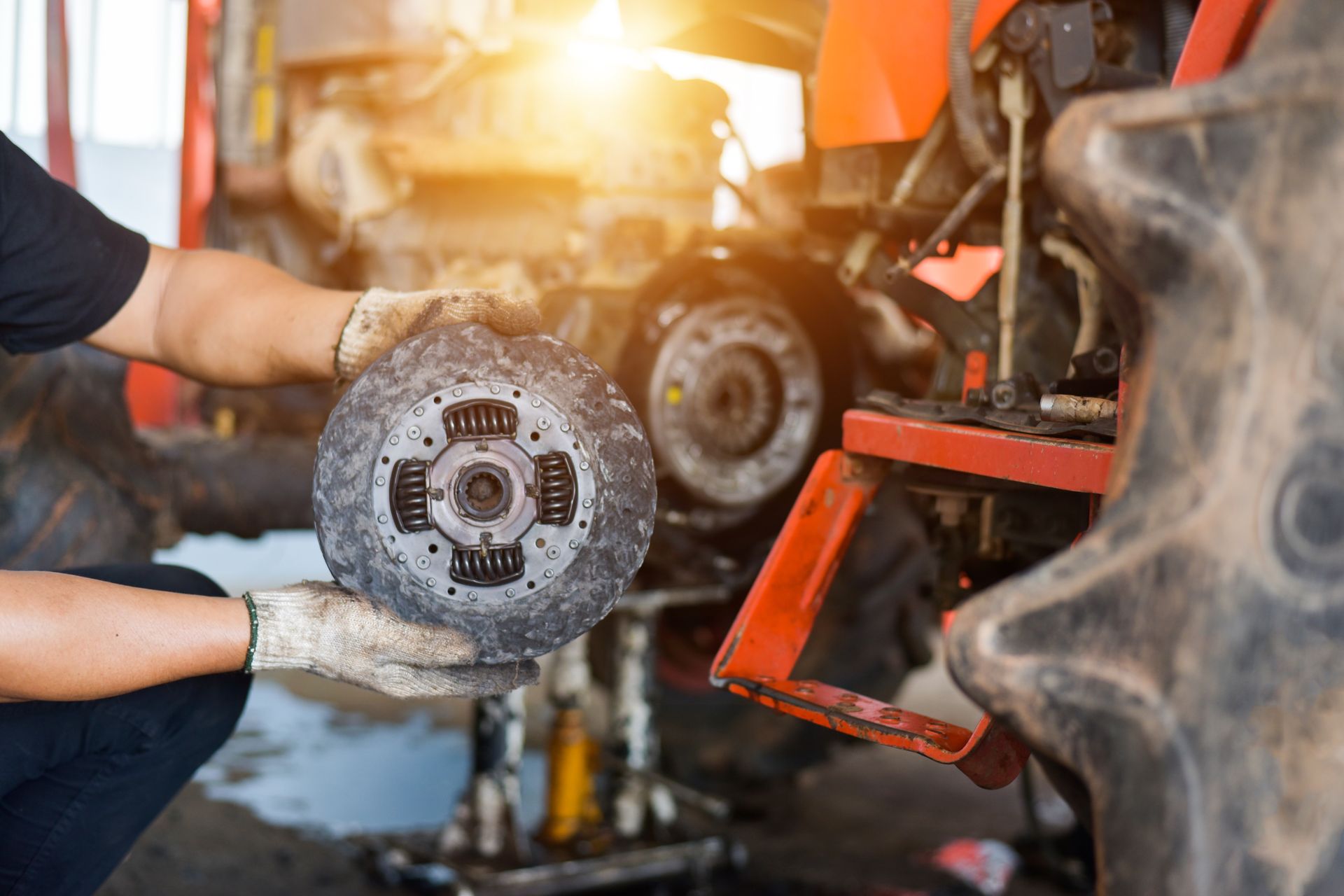 Mechanic holding a worn brake disc beside a tractor wheel in a workshop, with warm light in the background