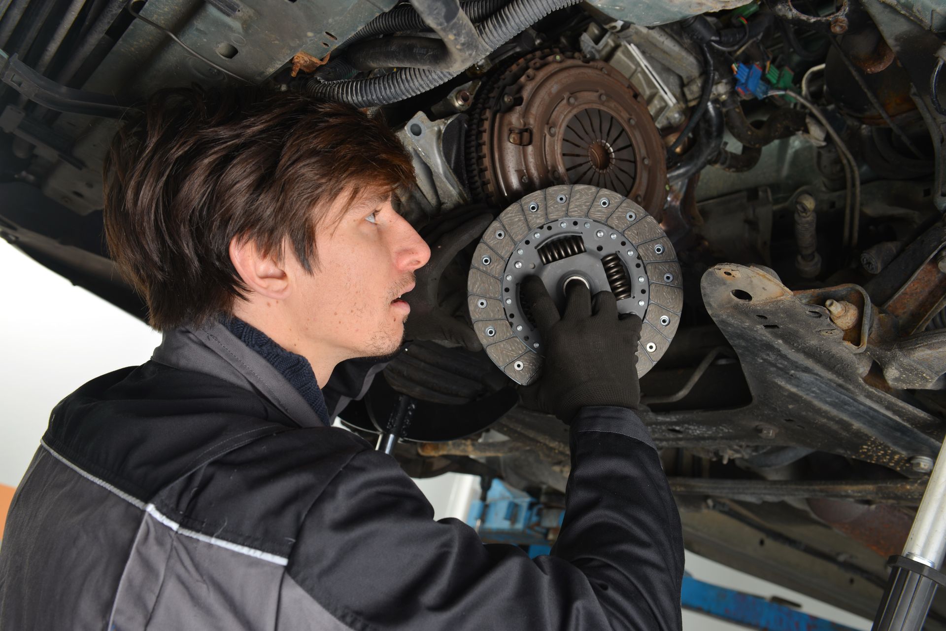 Mechanic inspecting a car brake assembly under a lifted vehicle, wearing gloves in a garage