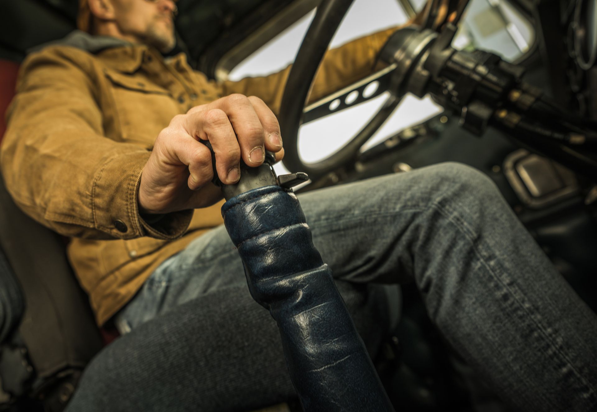 Driver’s hand on a gear shift inside a vehicle, with a tan jacket and jeans visible.