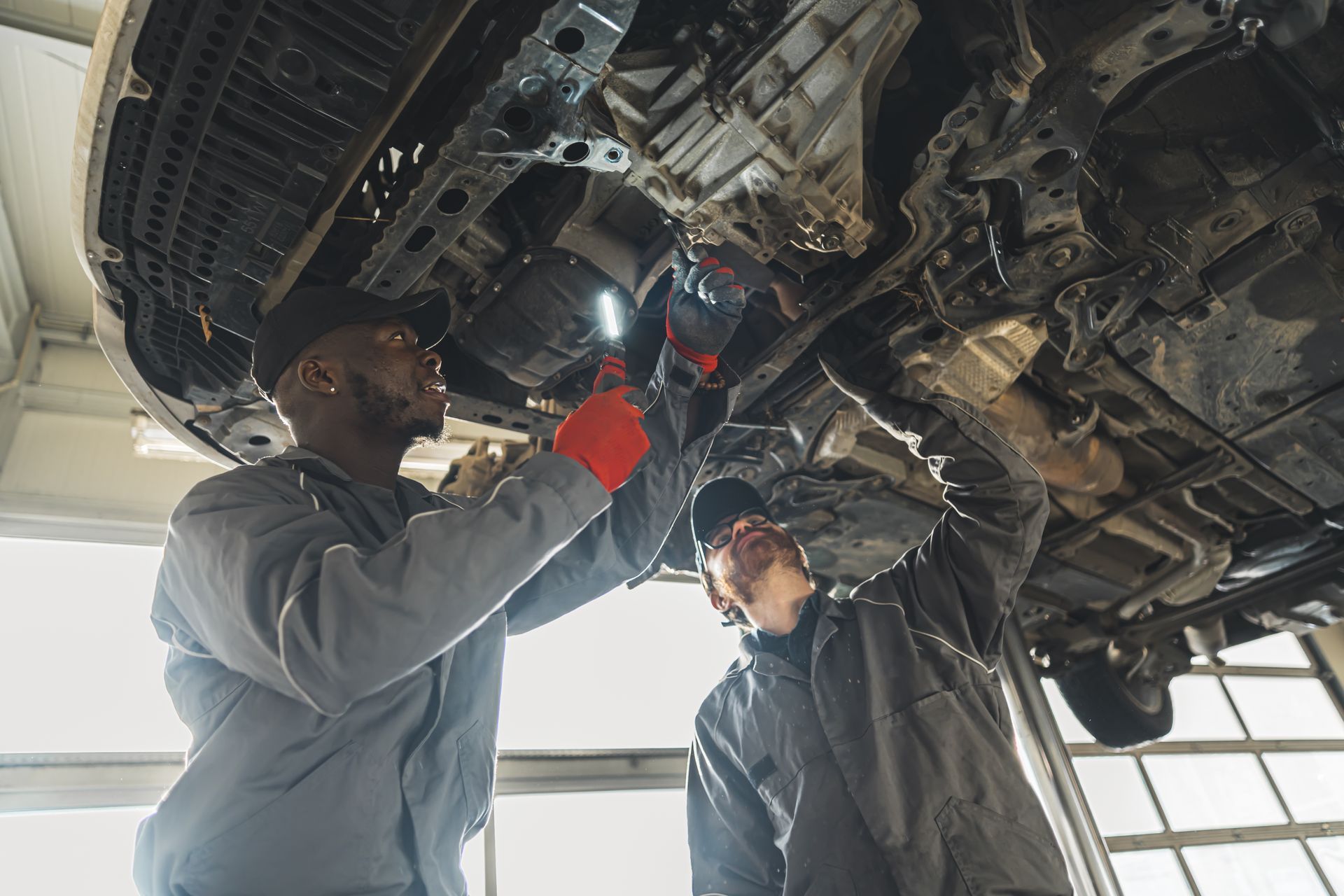 Two mechanics inspecting a car’s undercarriage in a garage, using a flashlight and tools.