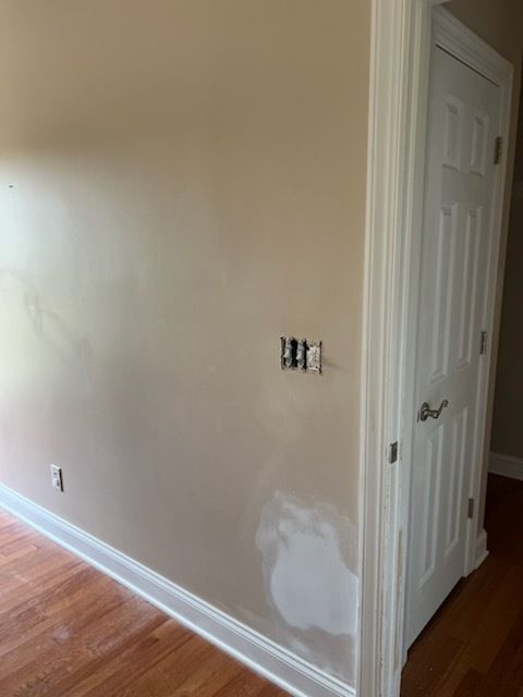 Empty beige hallway wall with exposed electrical box, white door, and patchy repair near baseboard.